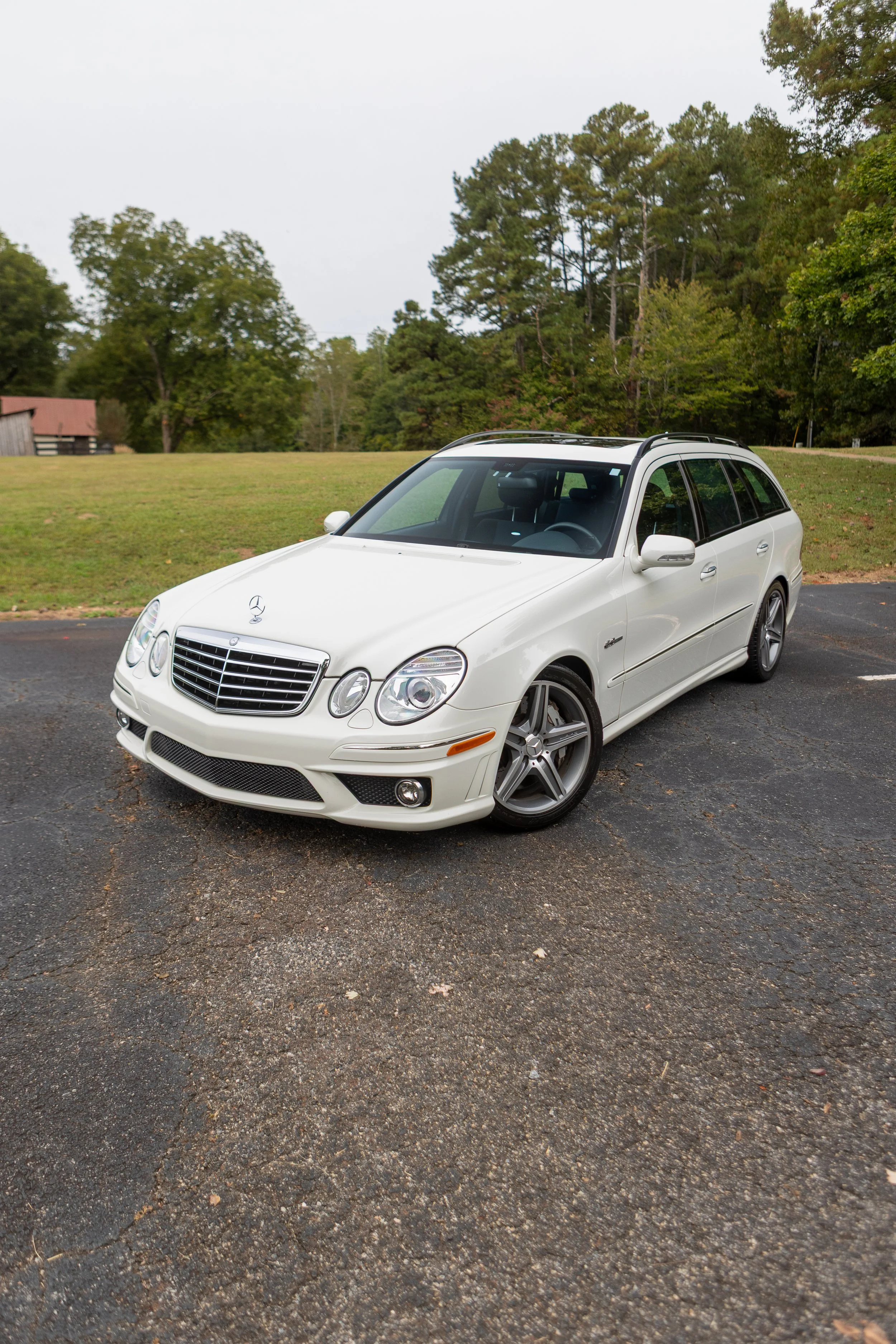 White Mercedes-Benz E-Class parked in a parking lot with green trees and a cloudy sky in the background.