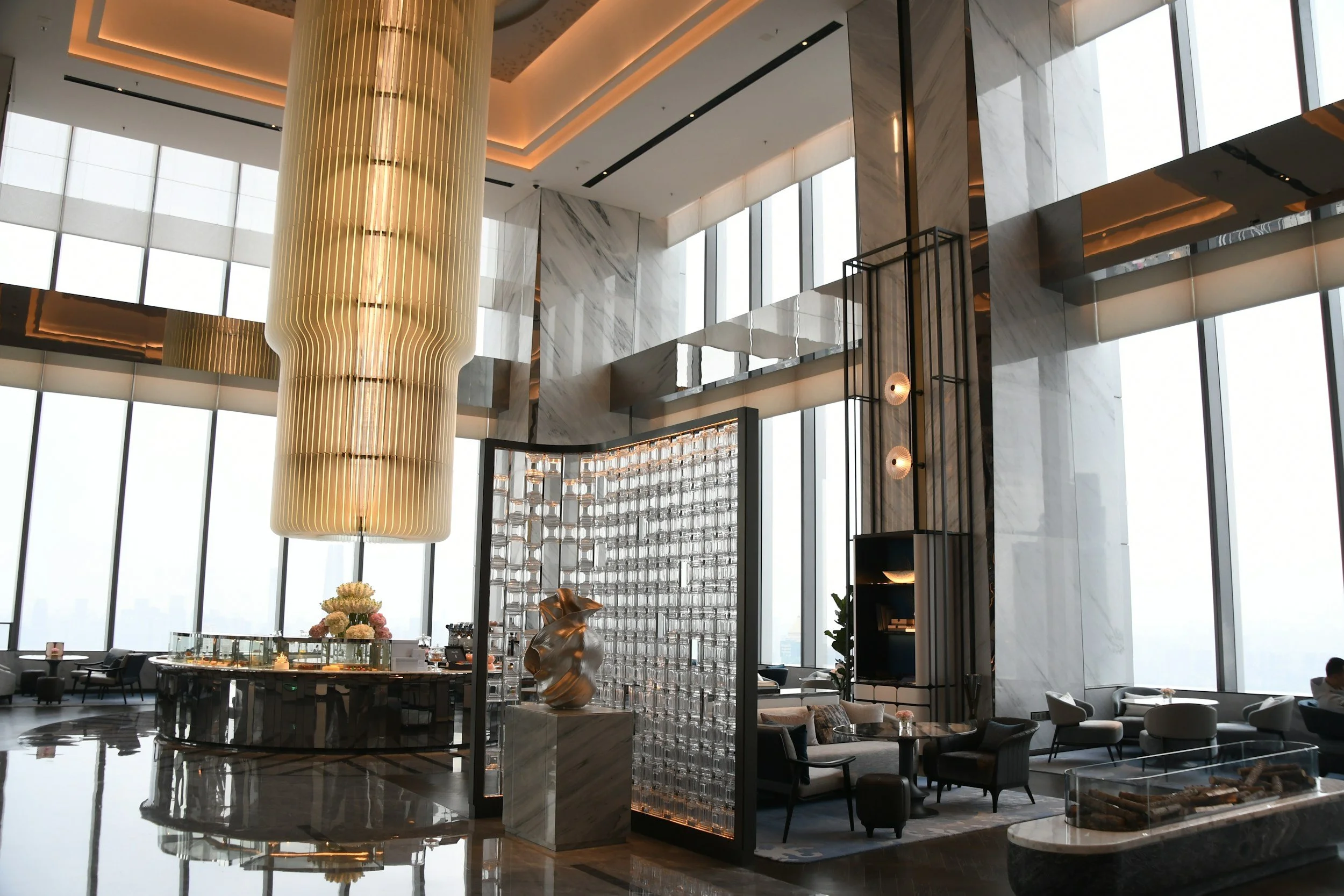 Interior of a modern high-rise building with large floor-to-ceiling windows, a tall vertical gold-colored chandelier, marble walls, and a seating area with tables and chairs.