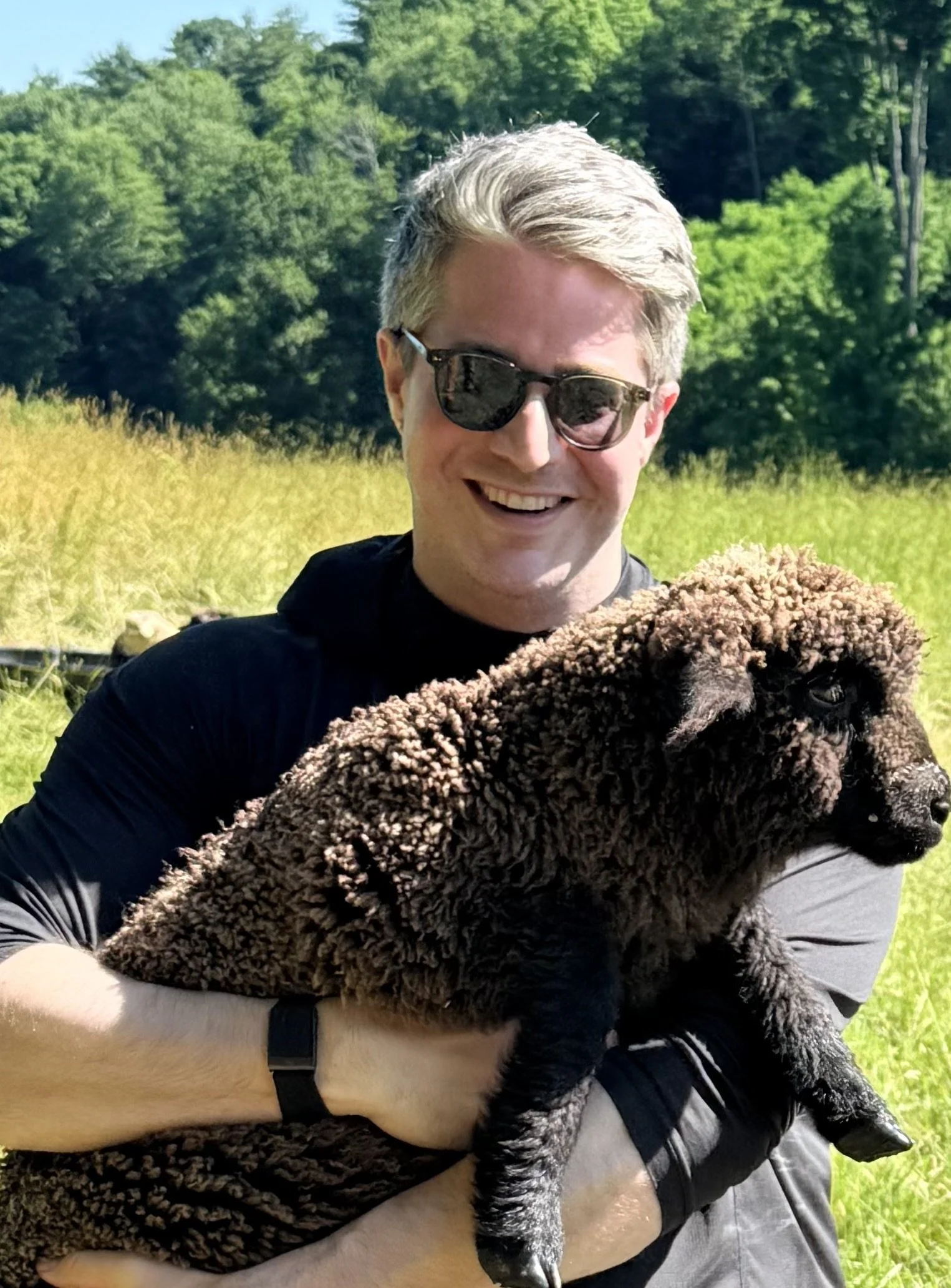 A person with blonde hair and sunglasses smiling and holding a large black lamb outside in a grassy field with trees in the background.