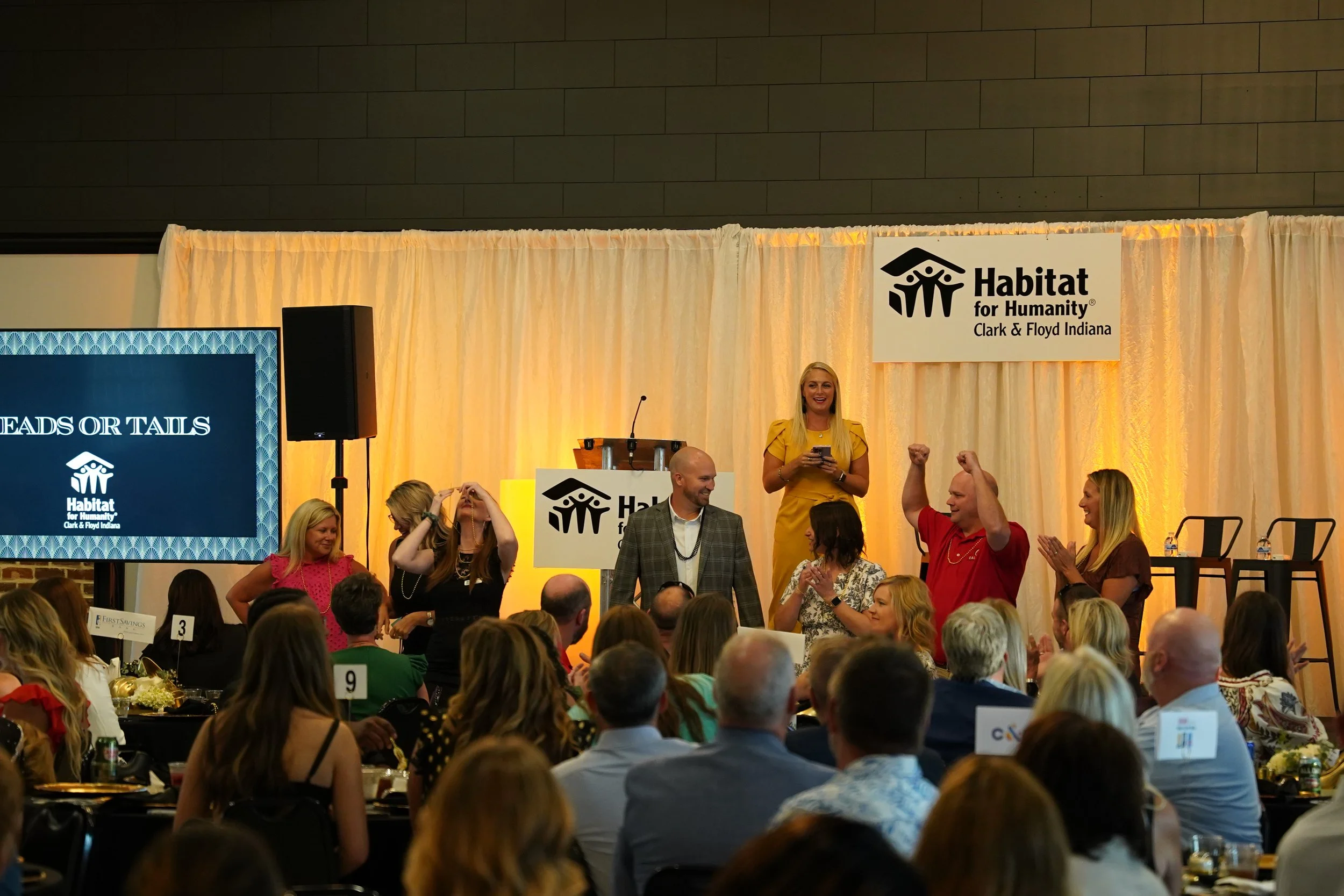 A large indoor event with many people seated at tables. A woman in a yellow dress is standing on stage smiling, while people around her cheer and raise their fists. A Habitat for Humanity banner is displayed on the stage backdrop, and a large screen shows part of the event name. People are engaged and smiling in the lively scene.