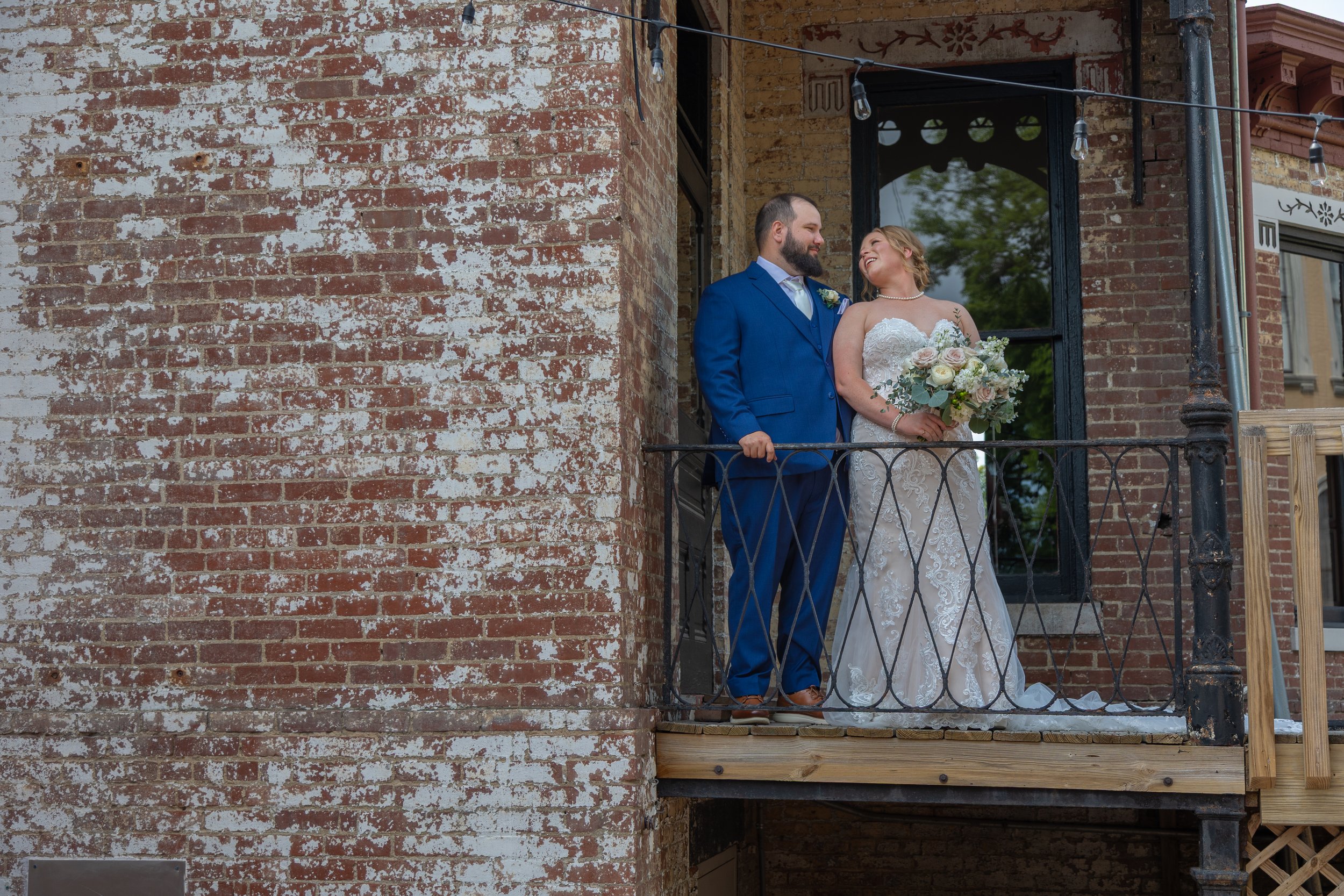 A bride and groom standing on a small balcony with black iron railing in front of a building with red and white brick wall. The groom has a beard and is wearing a blue suit, while the bride is wearing a white wedding dress and holding a bouquet of flowers.