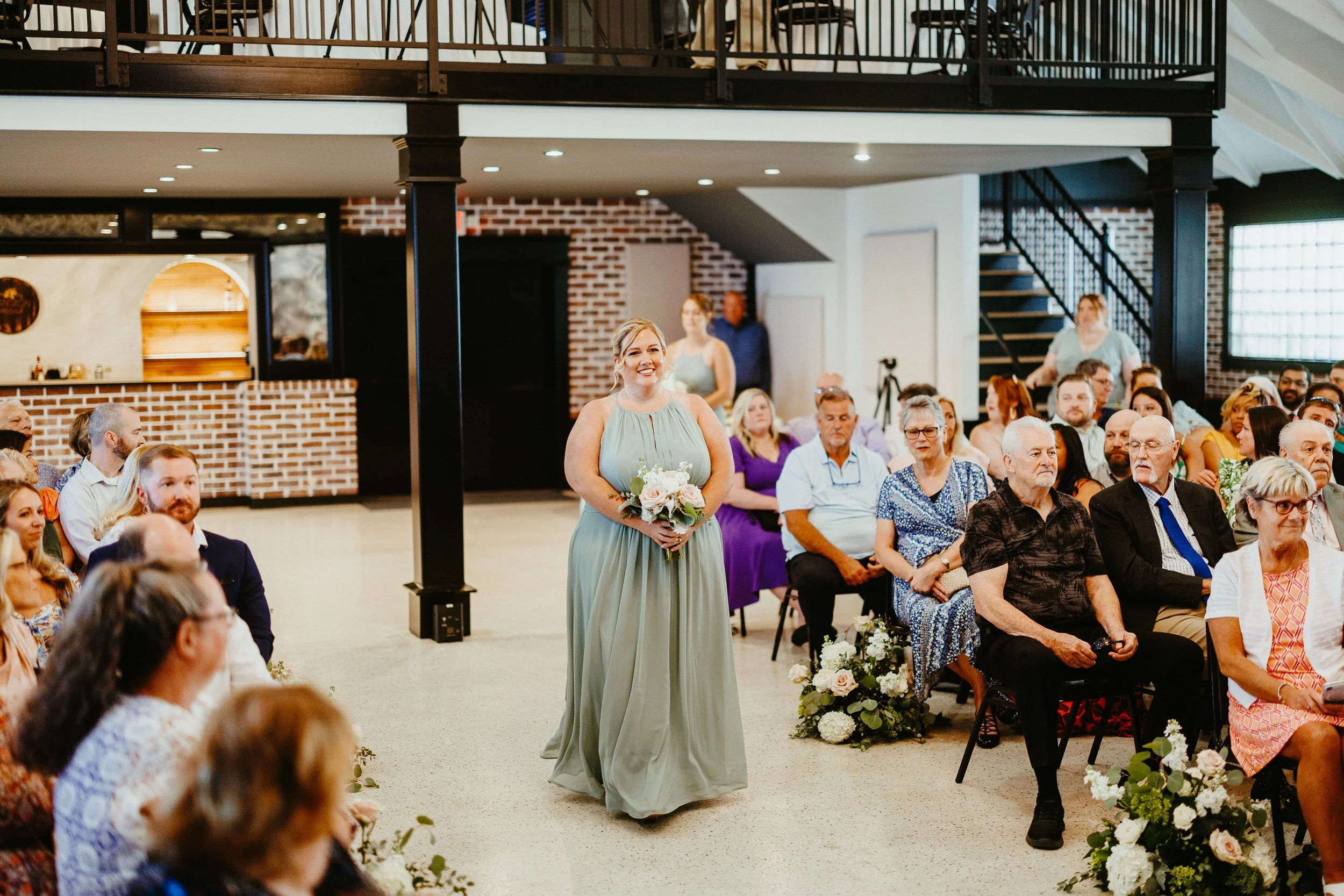 A woman in a light green dress holding a bouquet of flowers stands among seated people at an indoor wedding ceremony with exposed brick walls and modern decor.