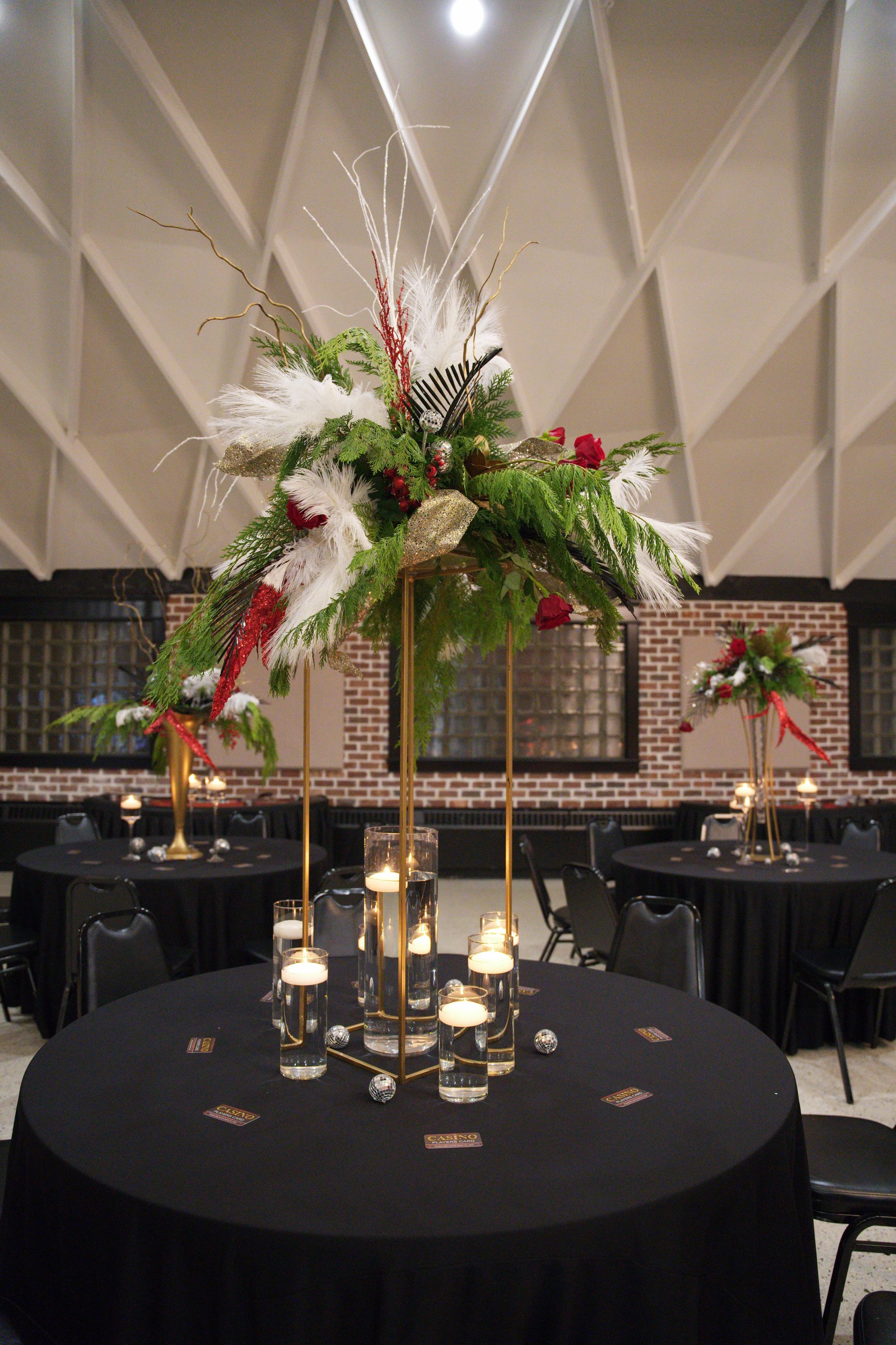 A large round table with a black tablecloth decorated with candles and a tall gold-colored centerpiece adorned with greenery, white feathers, red ribbons, and silver ornaments, set in a dimly lit event space with brick walls and additional similar tables in the background.