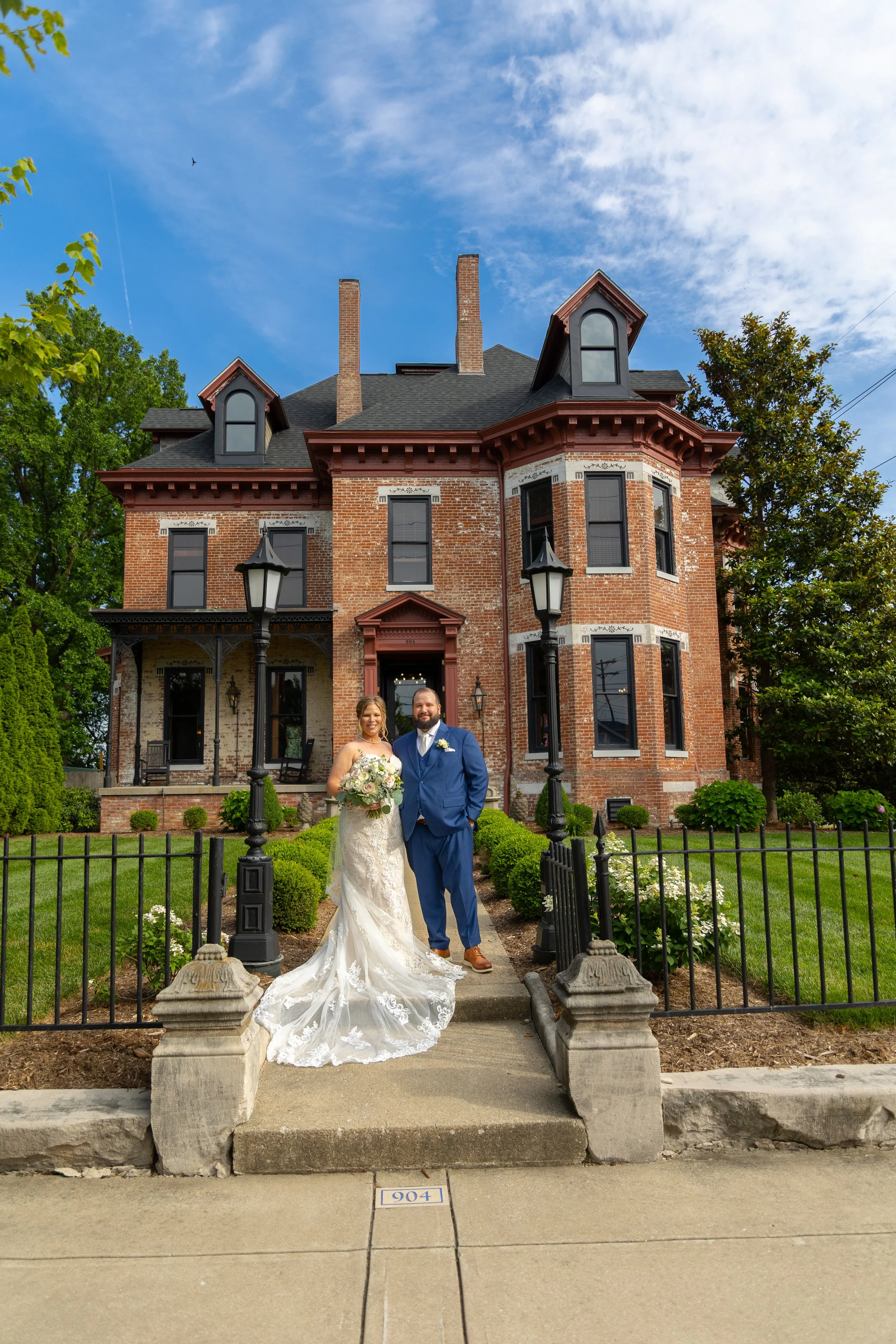 A bride and groom standing on a sidewalk in front of a historic brick house with a turret and arched windows, surrounded by greenery and black iron fencing, on a clear day with blue sky.