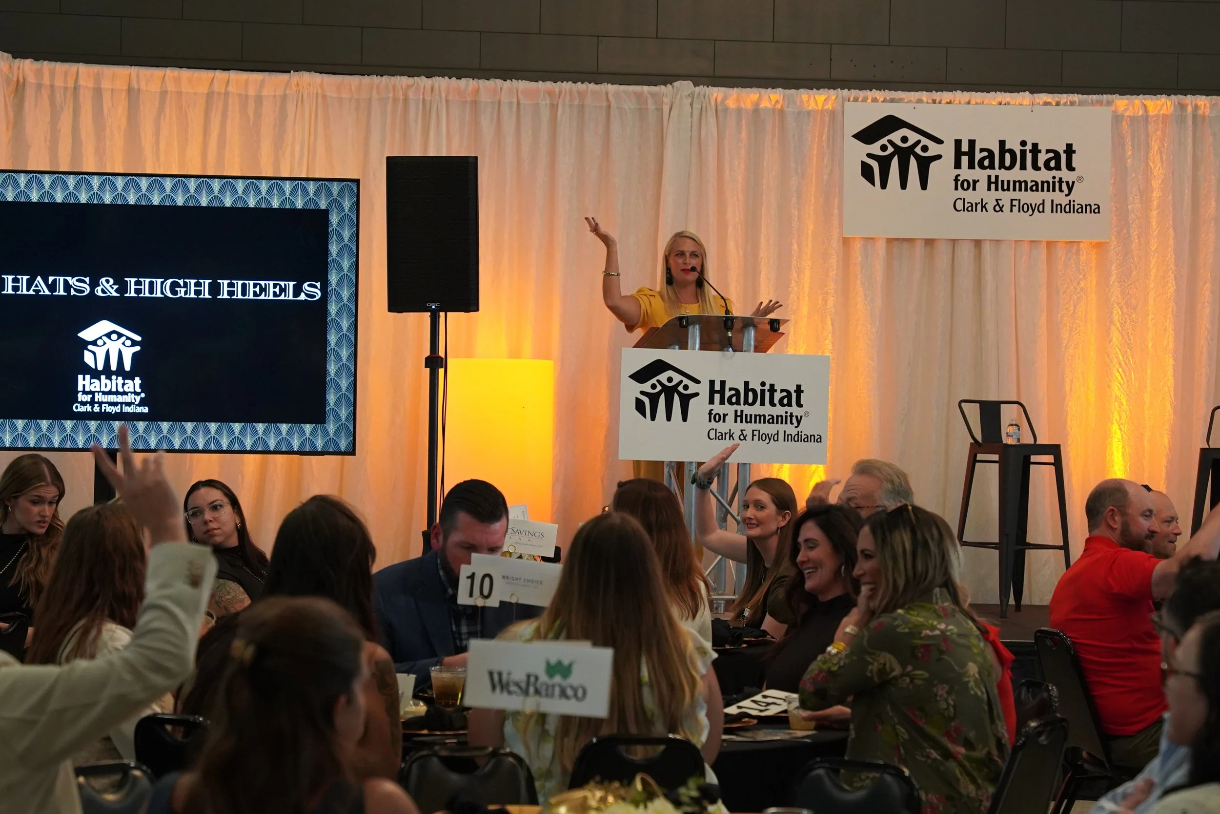 A woman with blonde hair wearing a yellow dress is speaking at a Habitat for Humanity event, standing behind a podium with a microphone. The background features a large Habitat for Humanity banner and a screen displaying the Habitat logo. Many attendees are seated at tables, some holding up signs with various names.