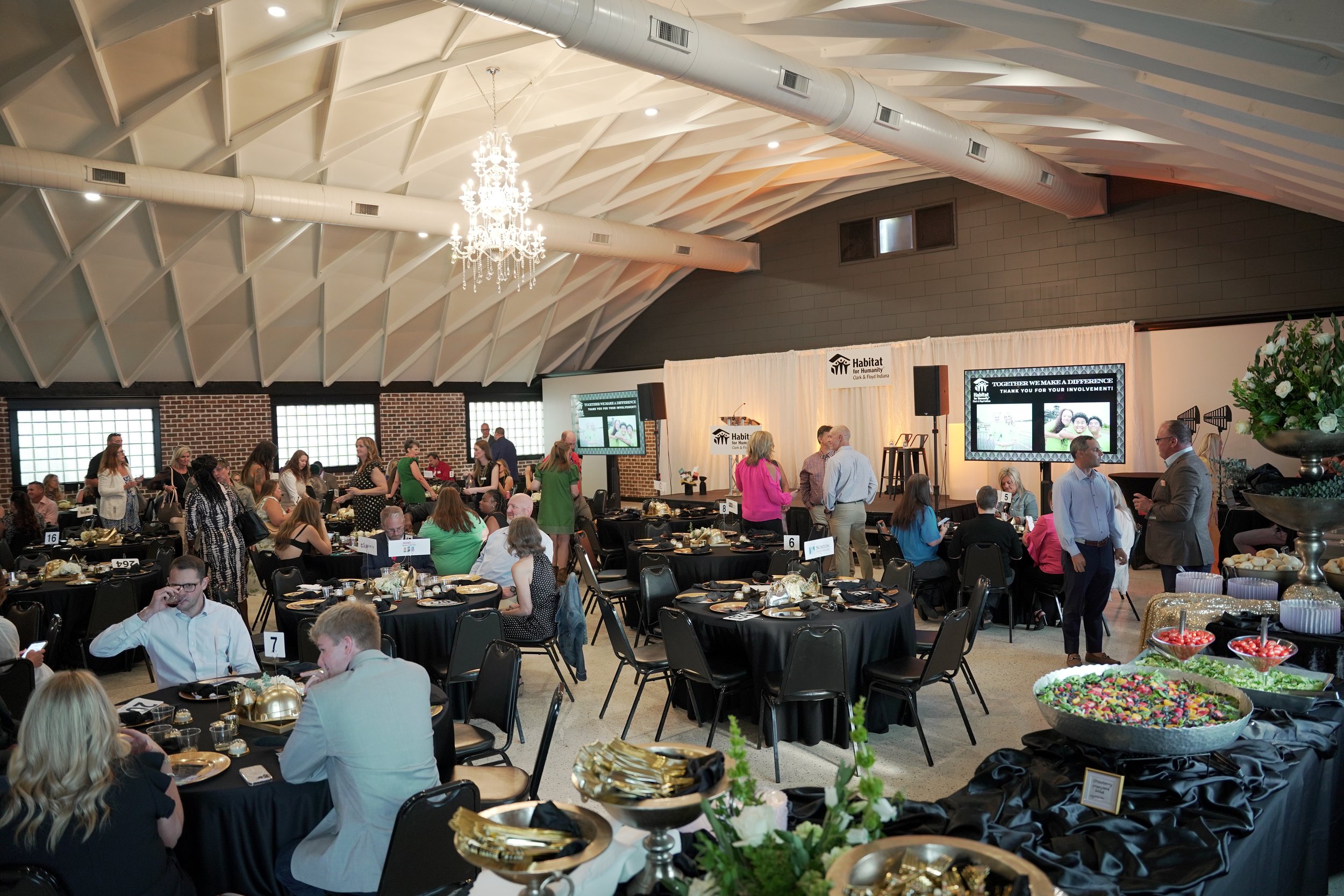 Indoor banquet hall with round tables set for a meal, decorated with gold and black tableware, and surrounded by people socializing. There is a large crystal chandelier hanging from the white vaulted ceiling with exposed beams and air ducts. In the background, a stage with a white curtain displays the