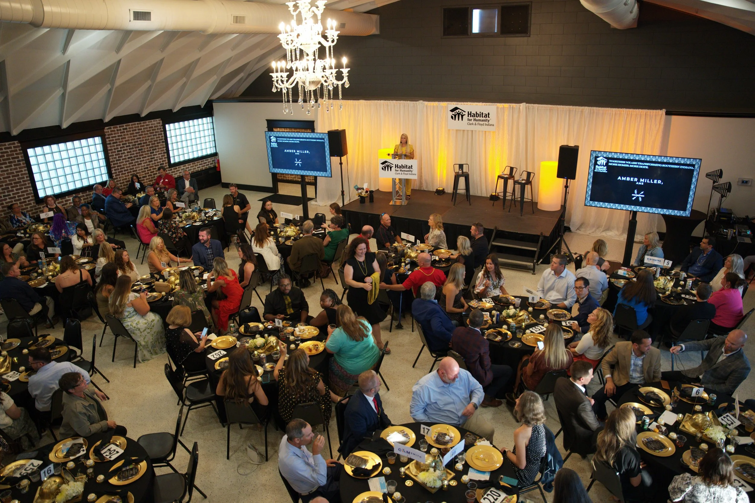 A large indoor event with many people seated at round tables decorated with black and gold table settings. A stage with a speaker at a podium and large screens displays the Habitat for Humanity logo. The room features a chandelier, brick walls, and several large windows.