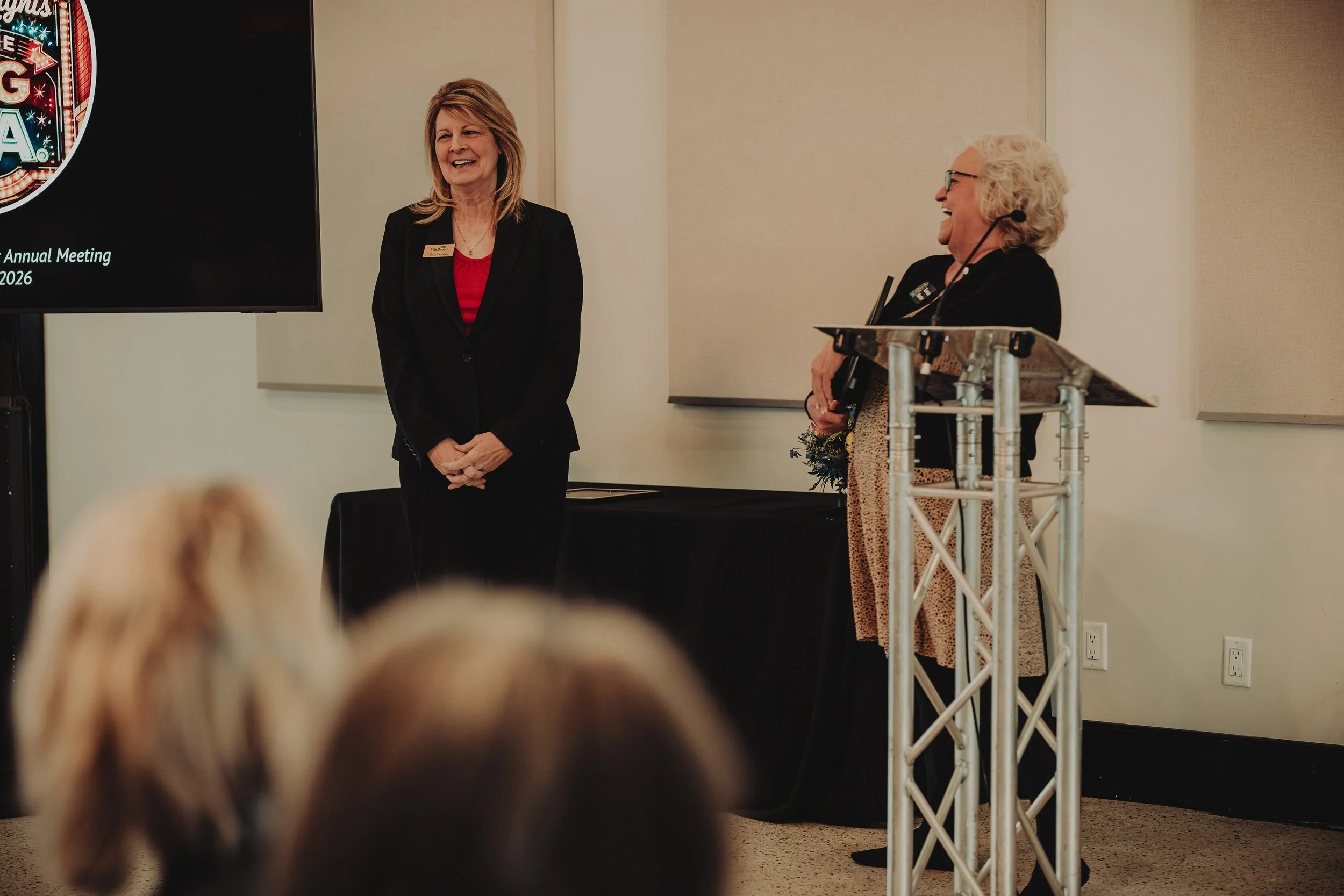Two women are standing at a podium and smiling during a meeting or event. One woman has shoulder-length blond hair and is dressed in a black jacket and red top. The other woman, with curly white hair and glasses, is wearing a black jacket and patterned dress, holding a bouquet of flowers. A large screen beside them displays event-related information.