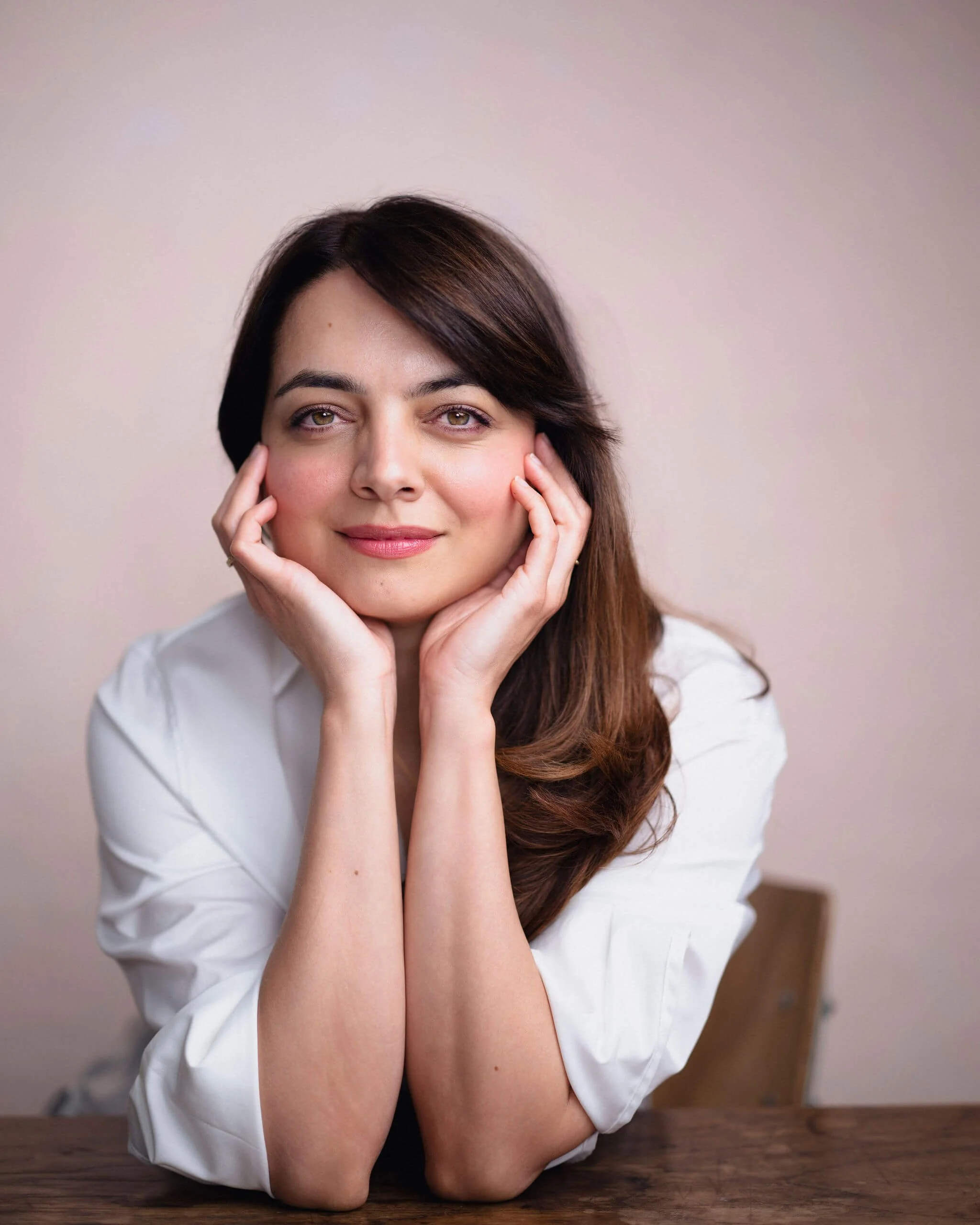 A woman with long brown hair and hazel eyes resting her face in her hands with elbows on a wooden table, wearing a white shirt, looking at the camera with a slight smile, against a plain beige background.