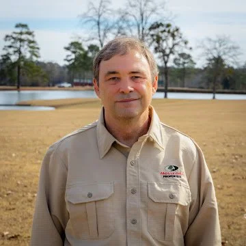 Man standing outdoors in a park with trees and a pond in the background, wearing a beige uniform shirt with a logo.