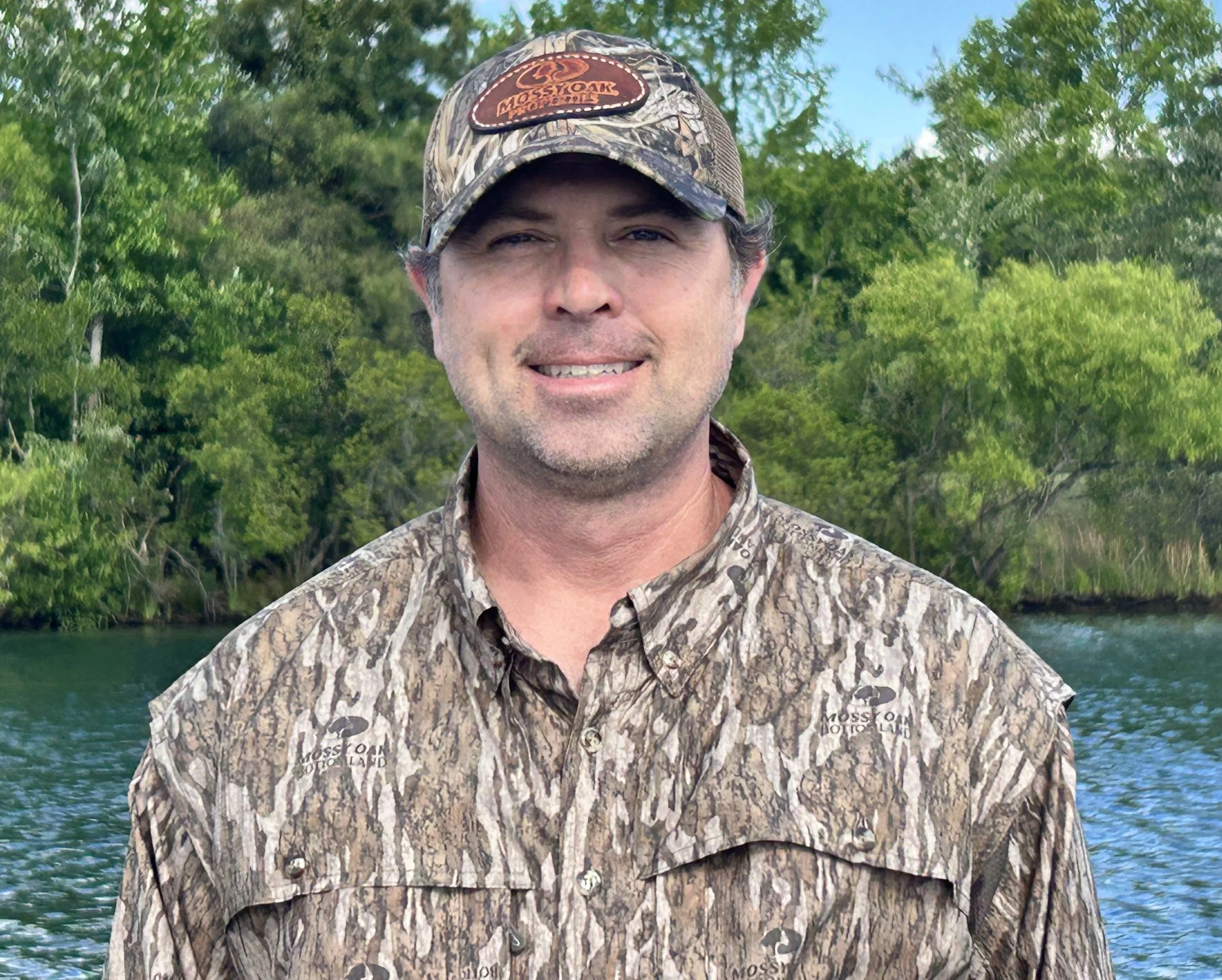 A man in camouflage clothing and a camouflage cap standing outdoors in front of a body of water and green trees.