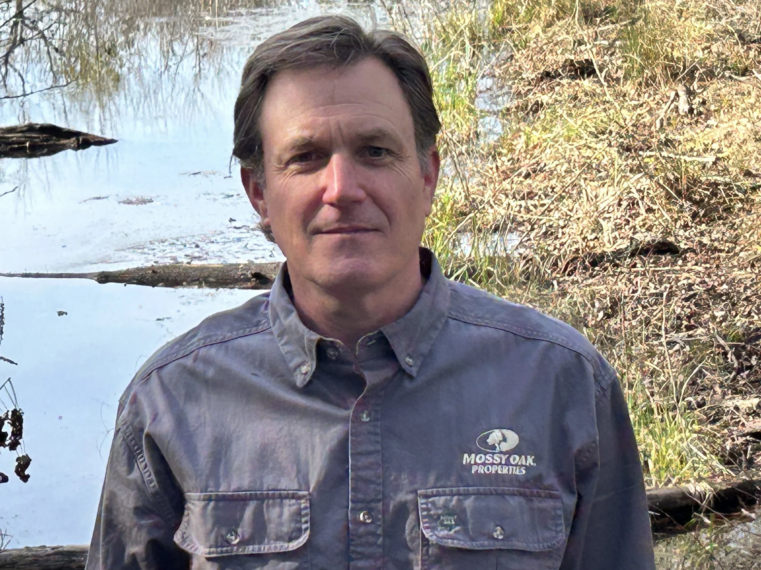 Portrait of a man standing in front of a natural pond or creek with trees and bushes around it. The man is wearing a Mossy Oak Properties jacket and looking at the camera.
