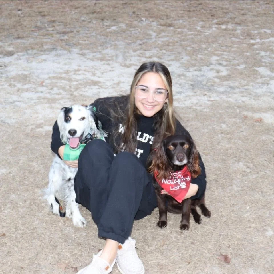 A young woman with long brown hair and glasses, sitting on the ground, smiling, with two dogs, a black-and-white spaniel and a brown spaniel, each wearing a red bandana, outdoors on a dirt or gravel surface.