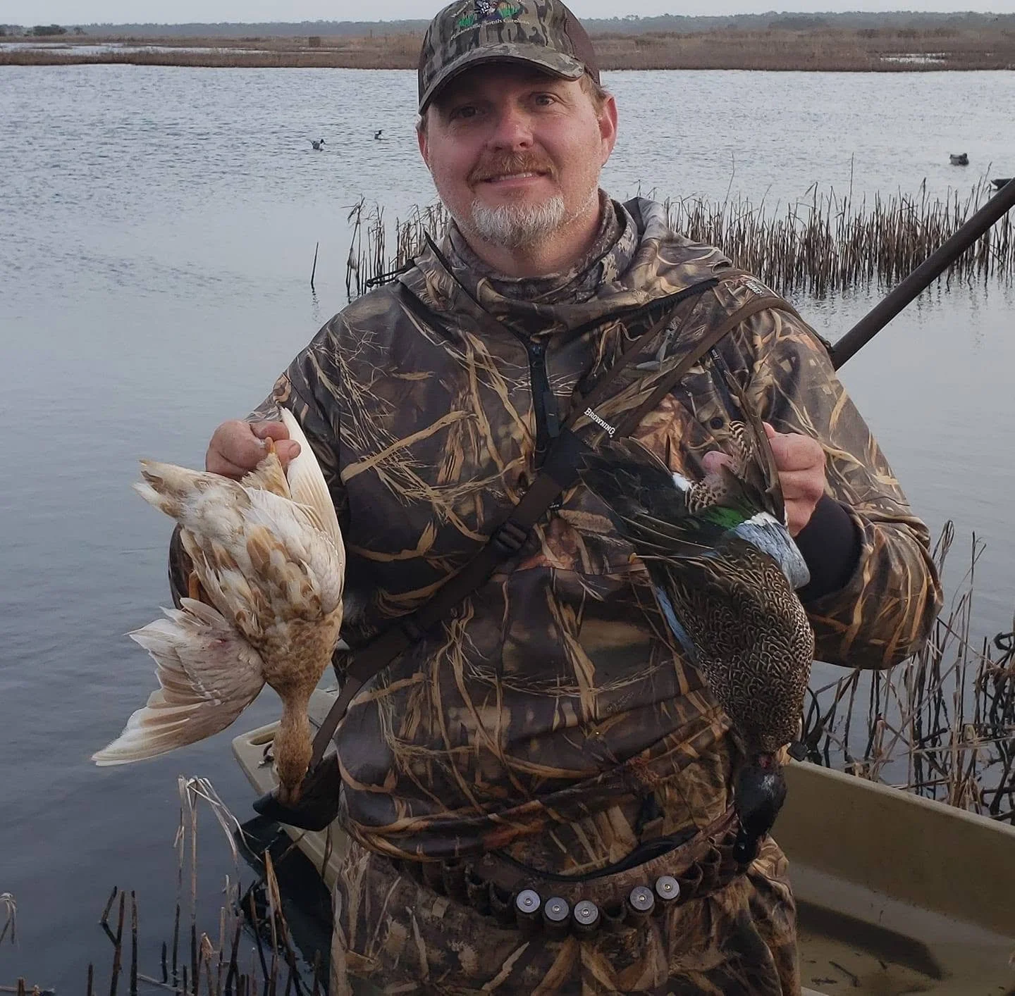 A man in camouflage clothing holding a duck and a goose near a lake with waterfowl, with reeds in the background.