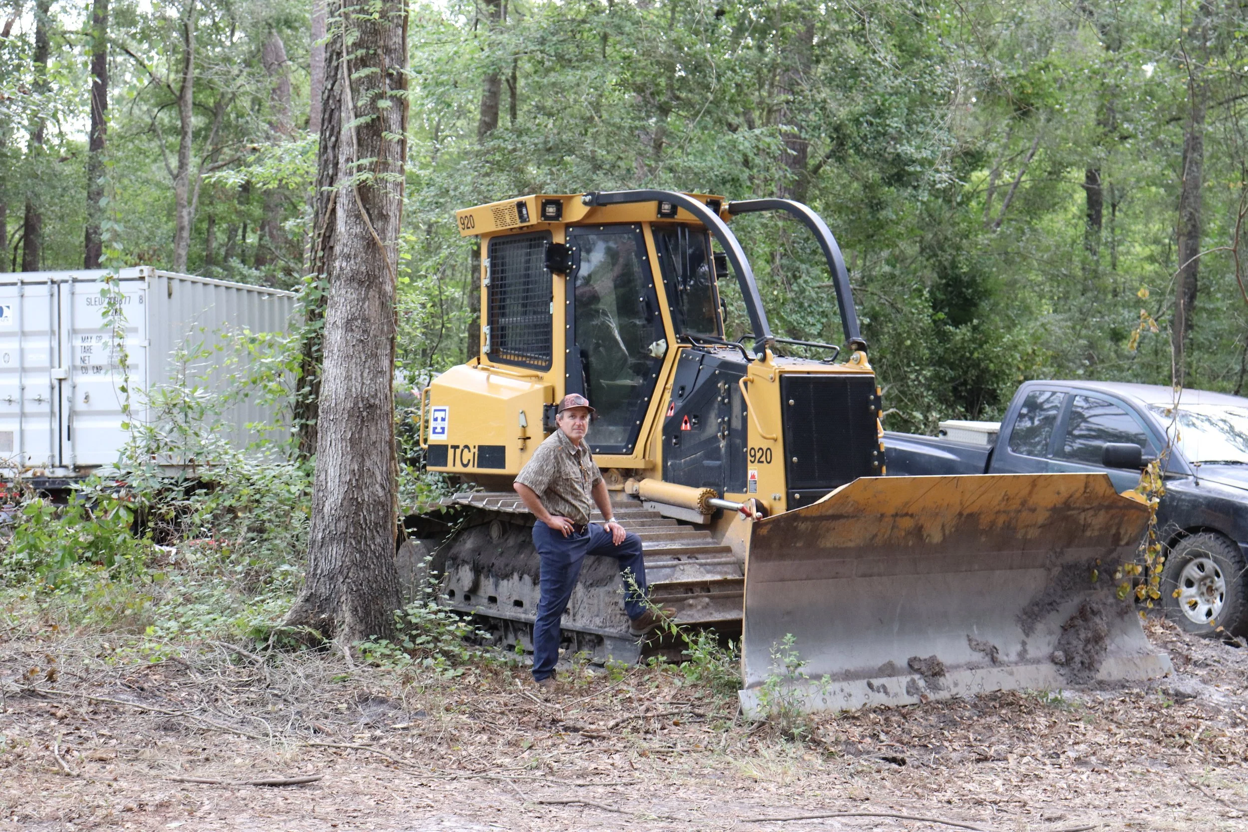 A person standing in front of a yellow bulldozer in a forested area with trees and foliage, alongside a black vehicle and a white container in the background.
