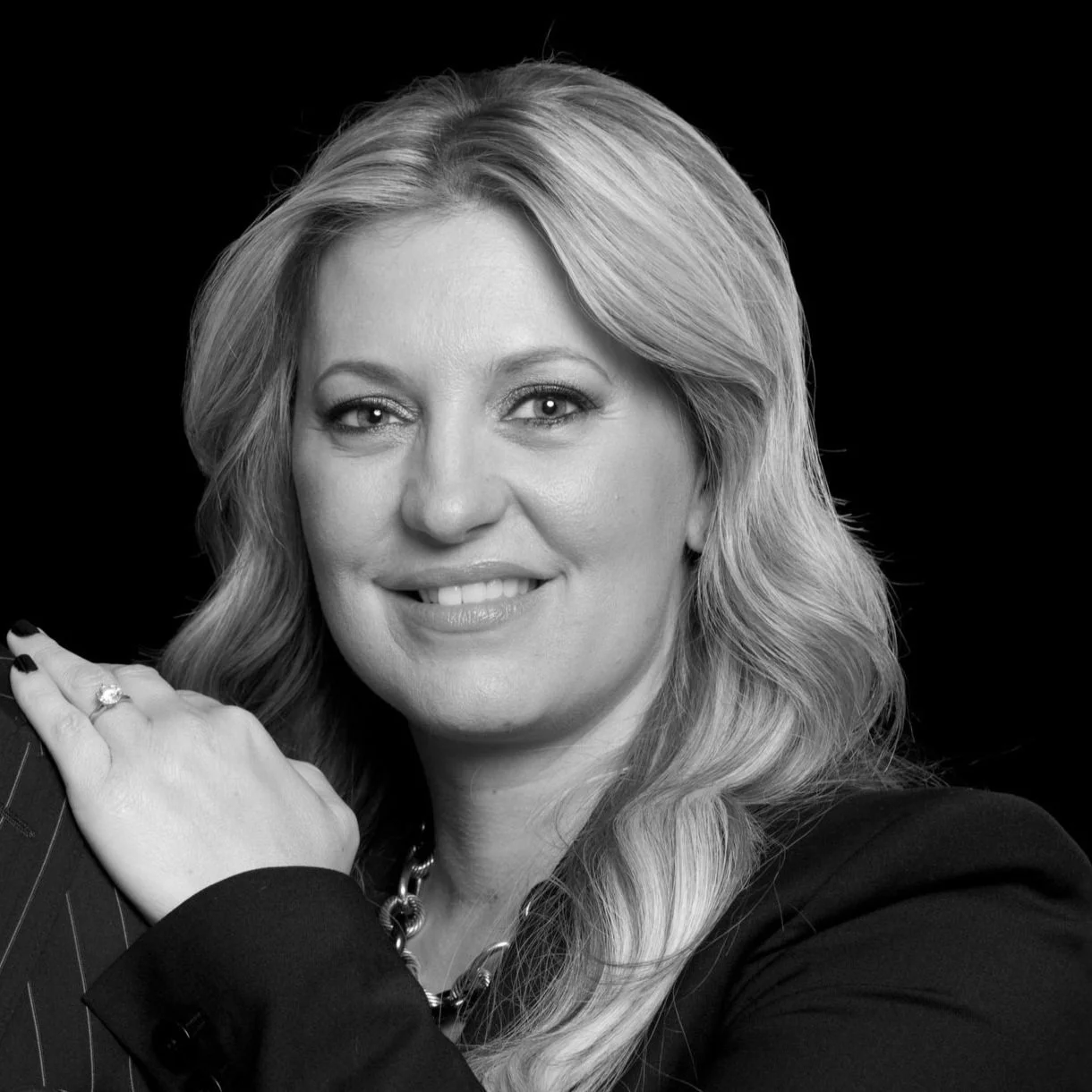 Black and white portrait of a woman with long wavy hair, wearing a dark blazer, necklace, and ring, smiling against a dark background.