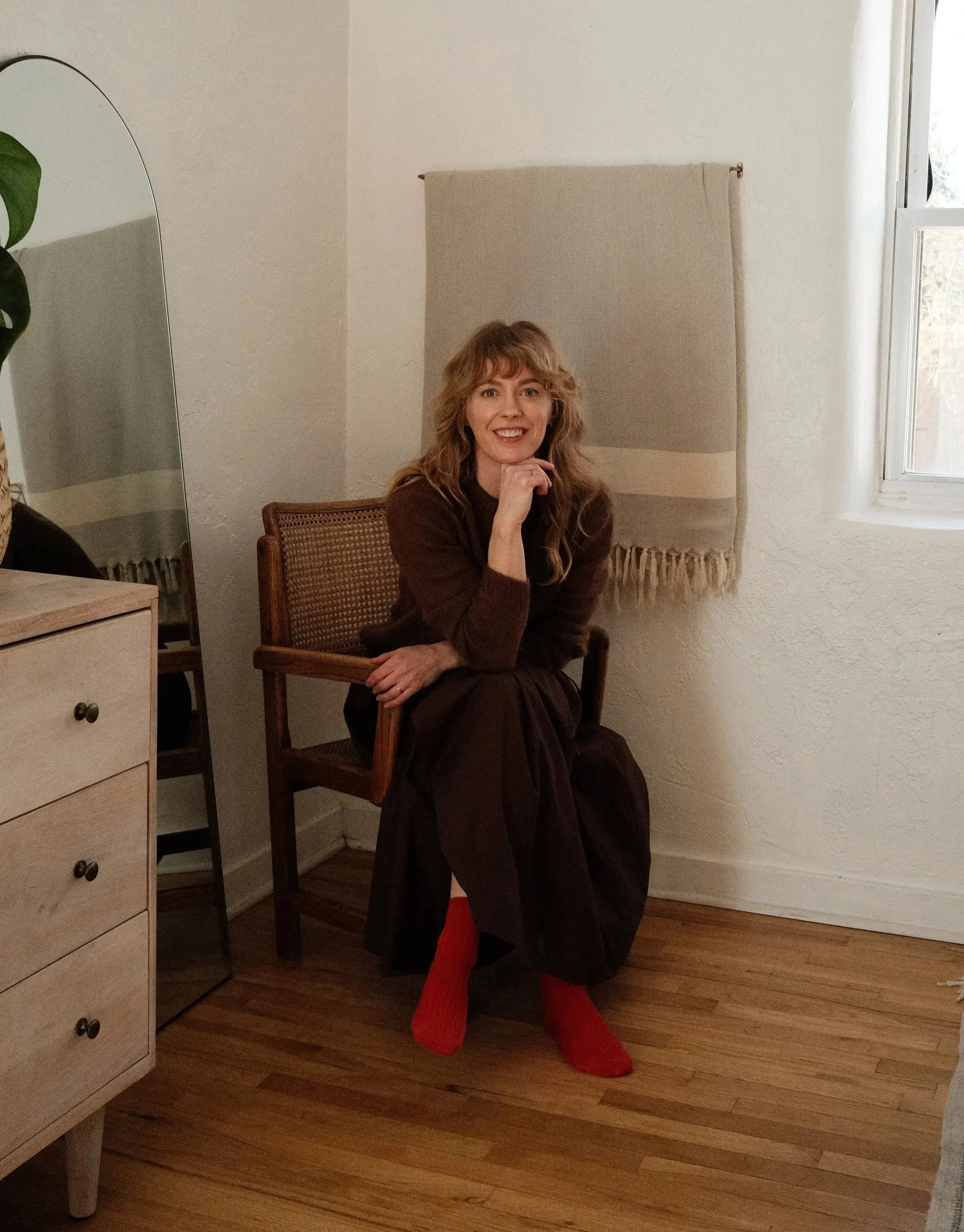 A woman sitting on a chair in a room with wooden floor, wearing red socks and a brown dress or coat, smiling and resting her chin on her hand. There is a white dresser to her left, a mirror, a beige wall hanging behind her, and a window on the right.