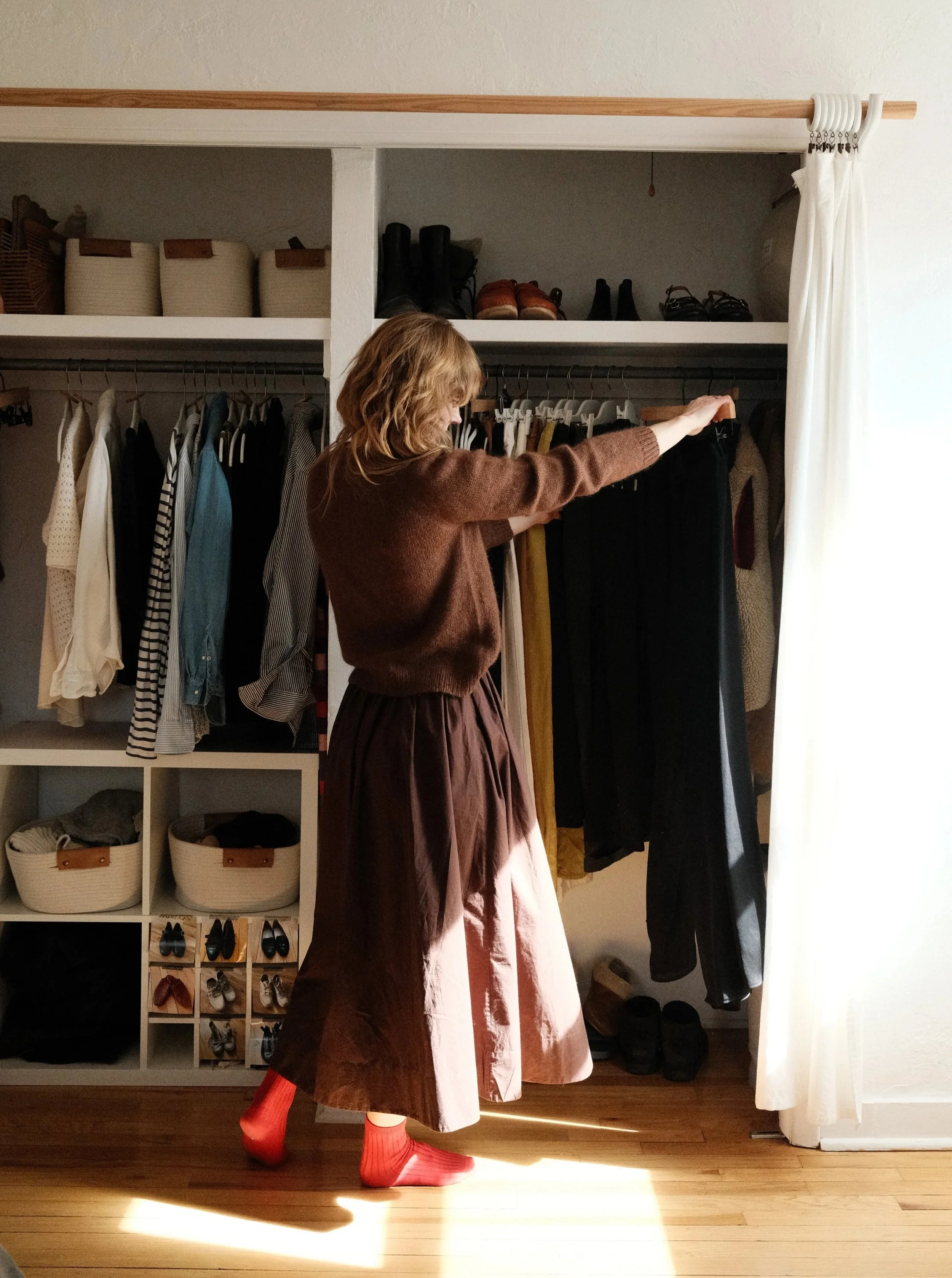 A person standing in front of an open closet with their head turned downward, wearing a brown sweater and a long skirt, with red socks visible. The closet has various clothes hanging, shoes stored in cubbies, and shoes on the floor. A curtain is partially pulled aside on the right side of the image.