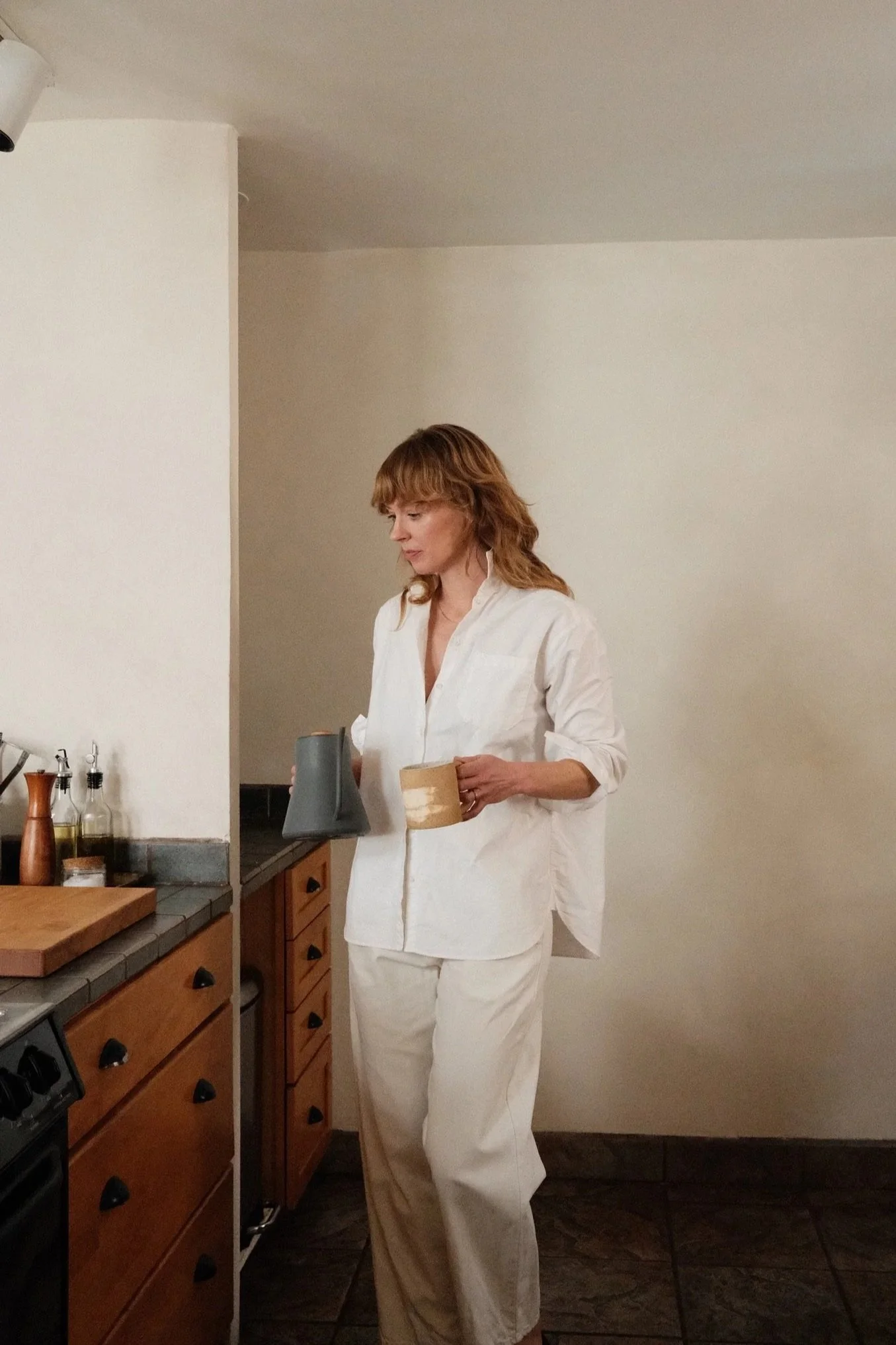 A woman wearing a white shirt and white pants standing in a kitchen, holding an object in her left hand, with wooden cabinets, kitchen utensils, and a countertop visible in the background.