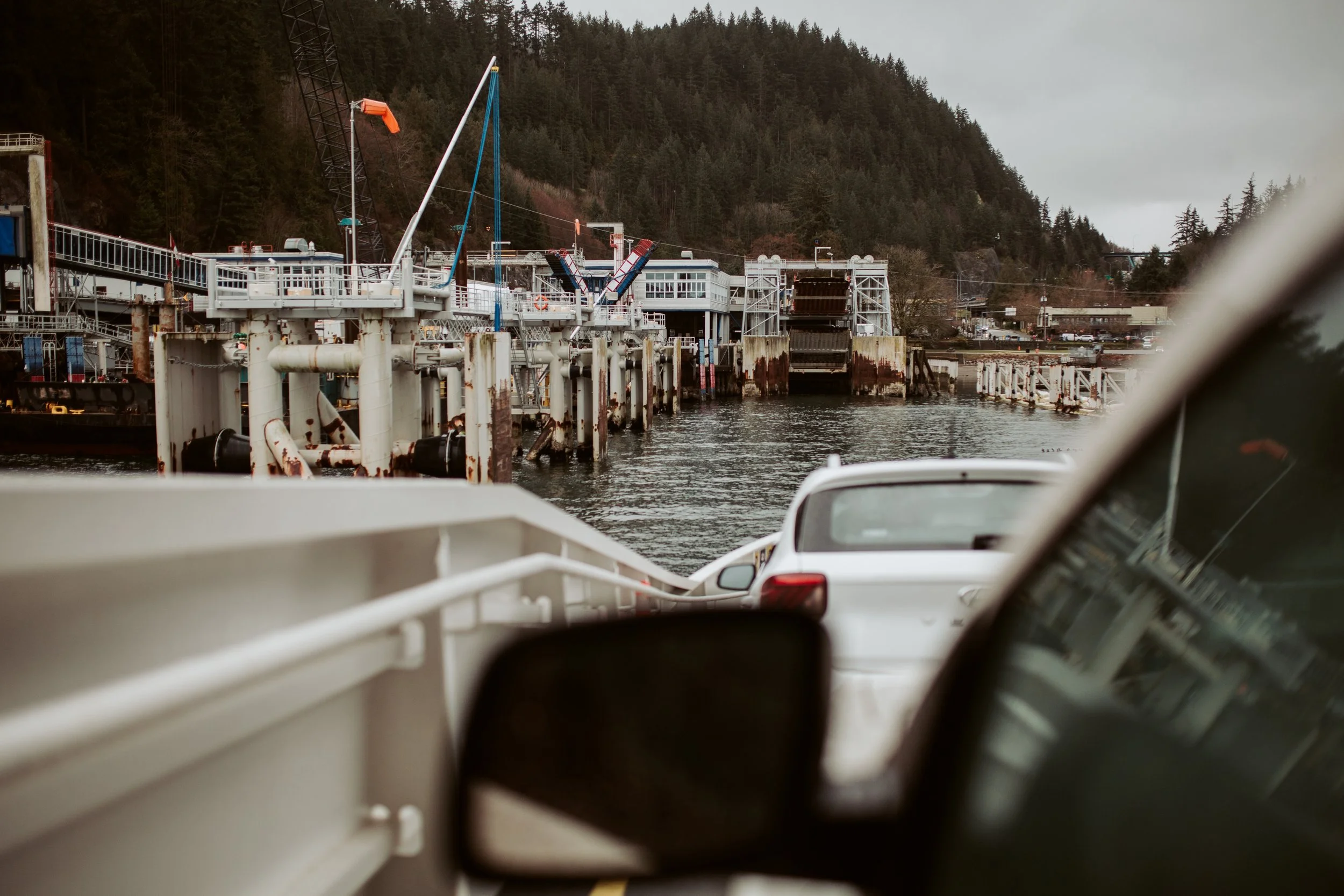 View of a dock area with boats, industrial structures, and vehicles, with forested hills in the background, overcast sky.