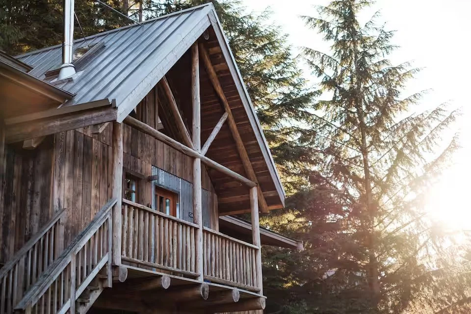 A wooden cabin with a steep metal roof, surrounded by tall pine trees, with sunlight filtering through the branches.
