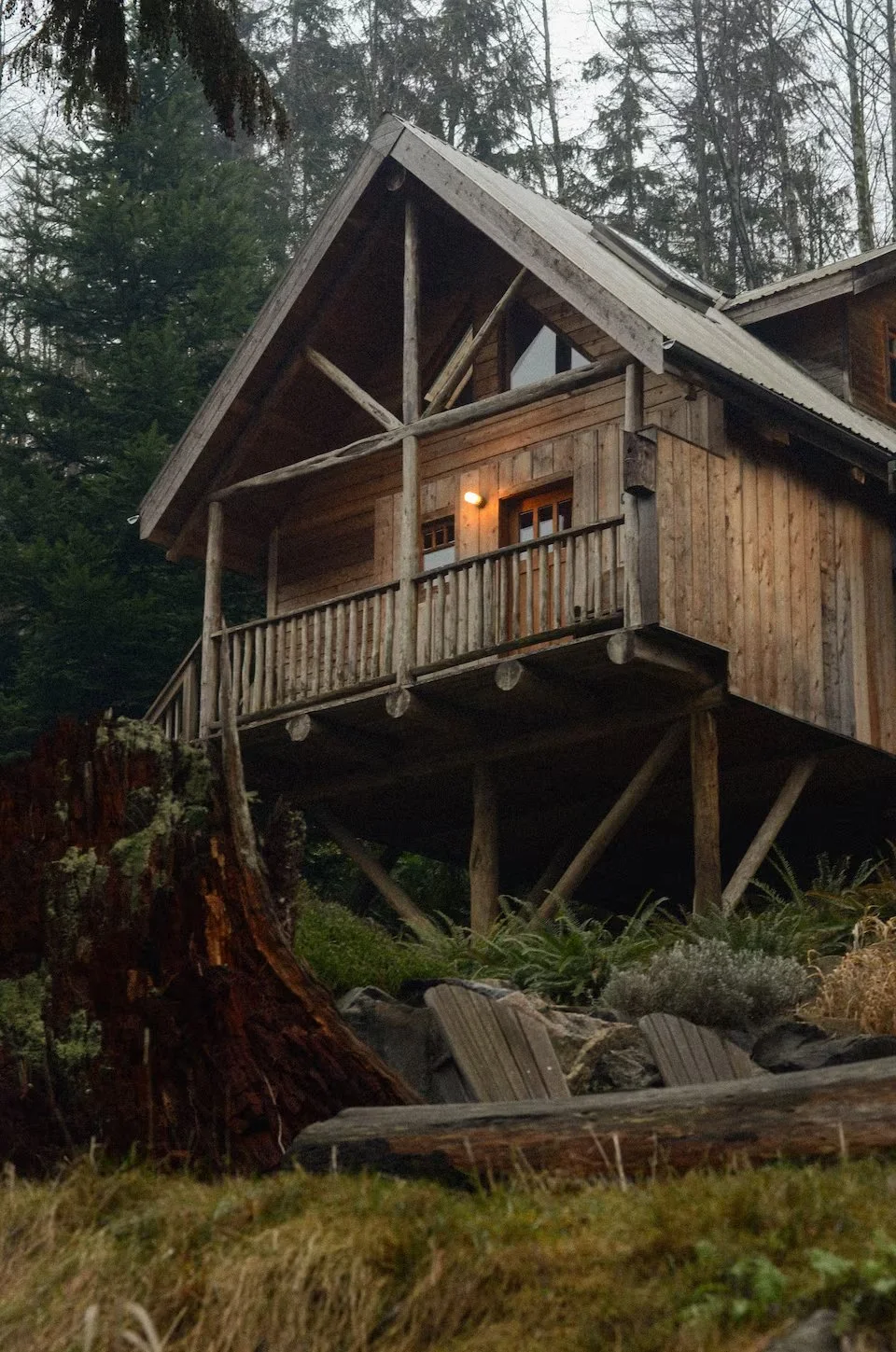 A rustic wooden house elevated on stilts in a forested area, with a small balcony, warm lighting, and a sloped roof.