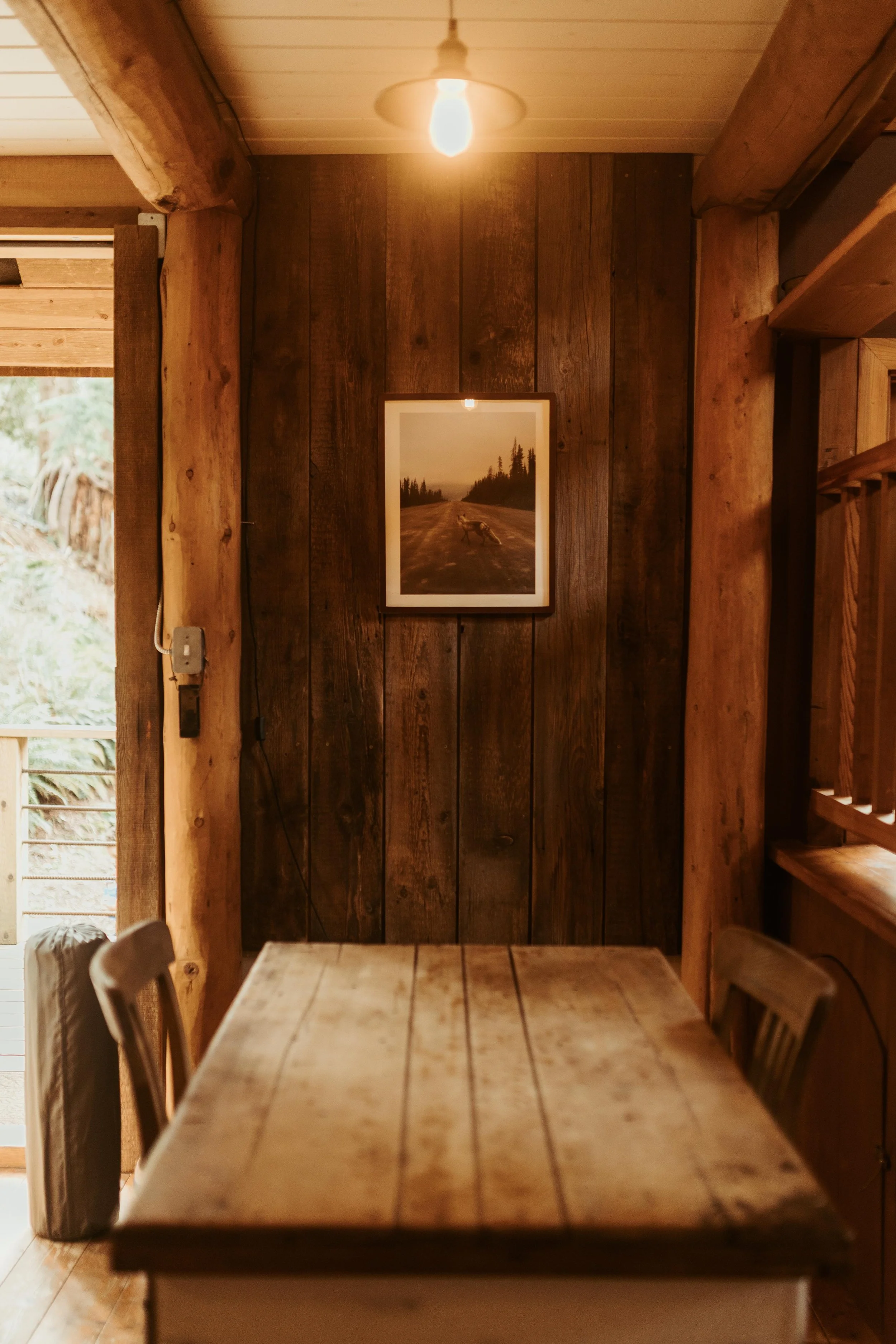 Interior of a rustic wooden cabin with a framed nature photograph on a dark wood wall, a dining table in the foreground, and a ceiling light bulb.