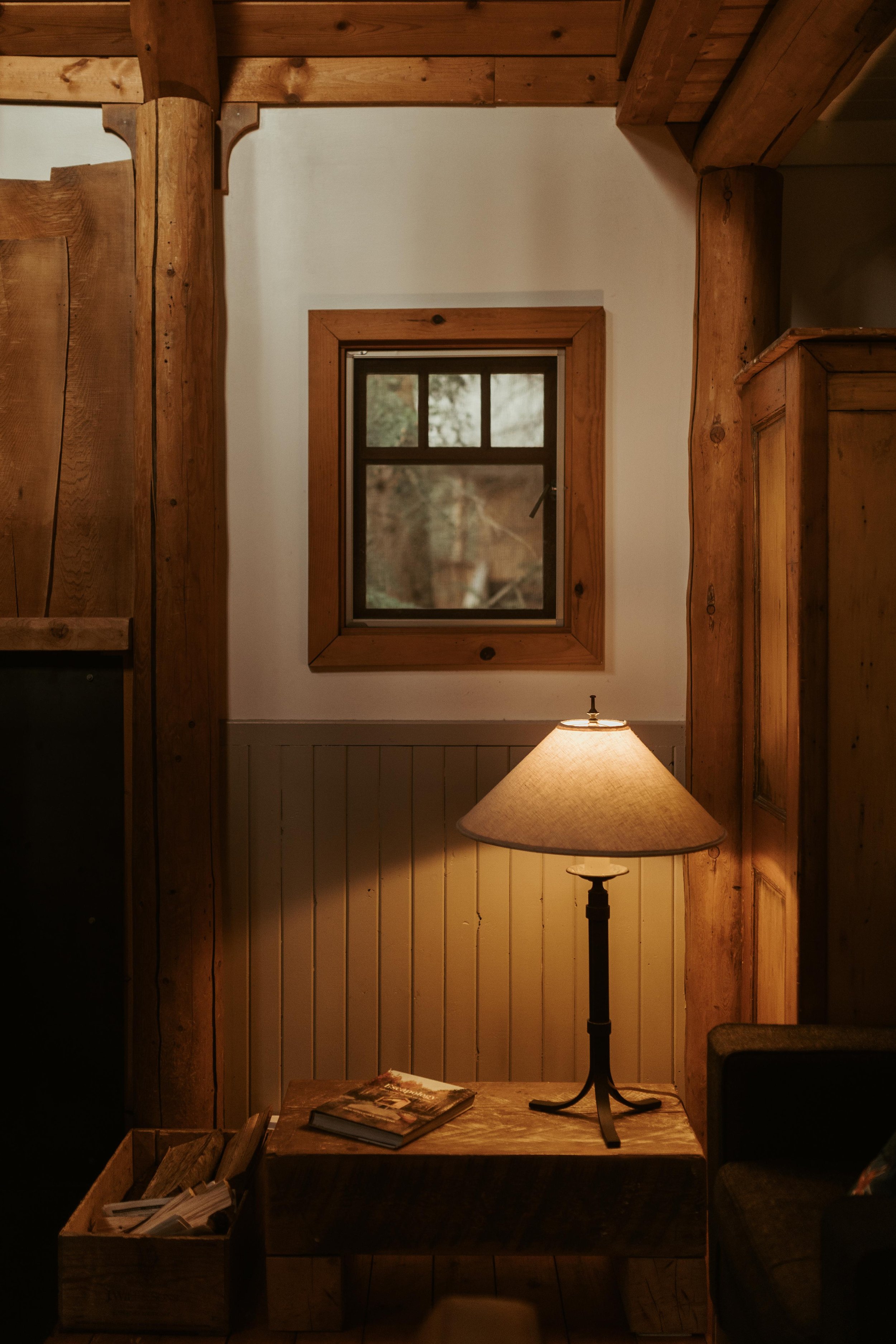 A cozy room with wooden beams and trim, a window on the white wall, a lamp on a table, books on the table, and a small bookshelf beneath the window.