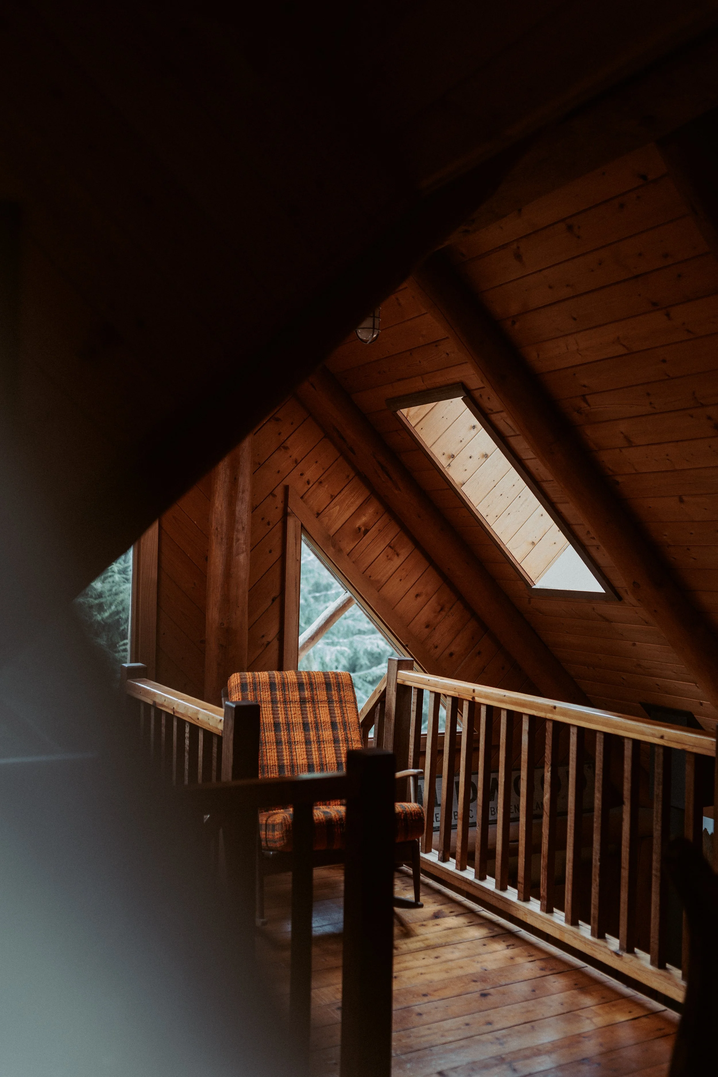 Interior of a wooden cabin with sloped ceiling and skylight windows, featuring a plaid upholstered chair, wooden flooring, and railing.