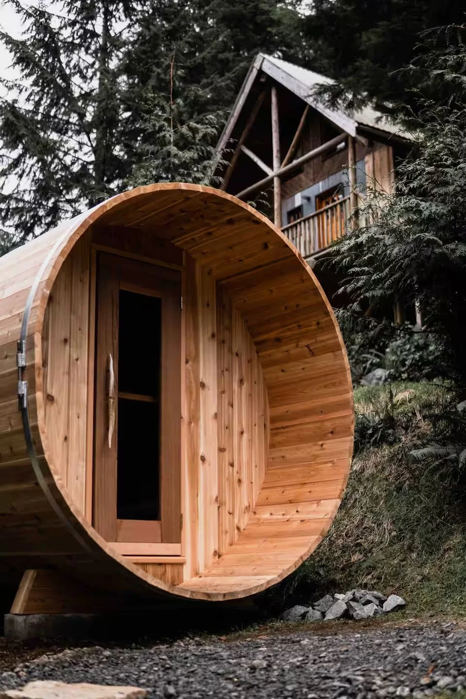 A wooden barrel-shaped small house with a door, situated outdoors among trees, with another similar house on stilts visible in the background.