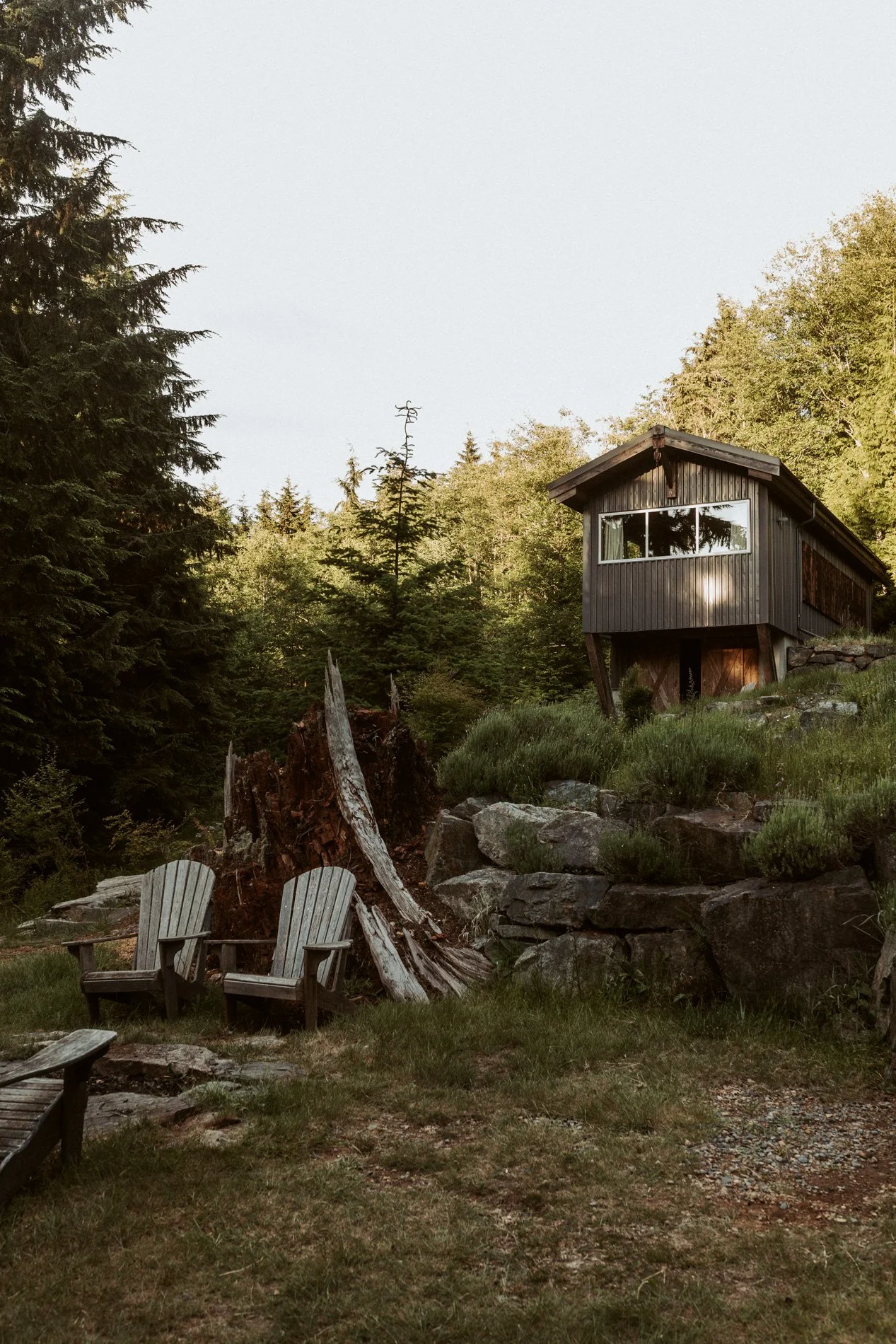 A house on stilts in a forested area with trees and rocks, and two Adirondack chairs on a grassy patch in the foreground.