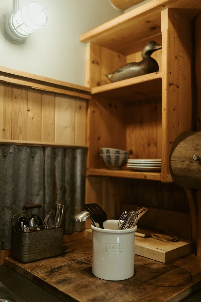 Kitchen countertop with utensils in a white container, small metal basket, wooden shelves with a duck figurine, bowls, and plates in a rustic kitchen setting.