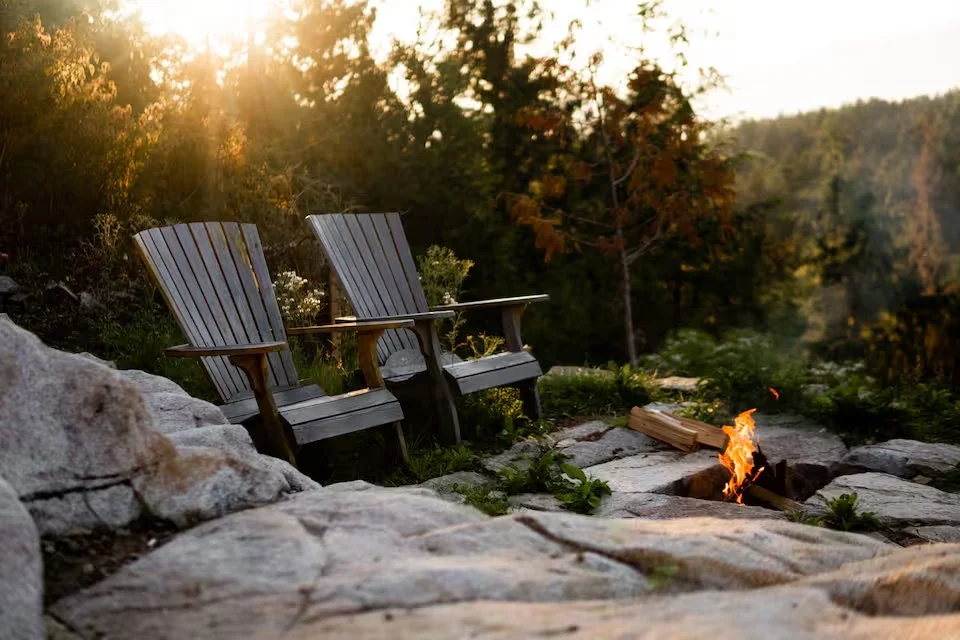 Two wooden Adirondack chairs sitting on a rocky area near a small campfire, surrounded by trees and foliage during sunset.