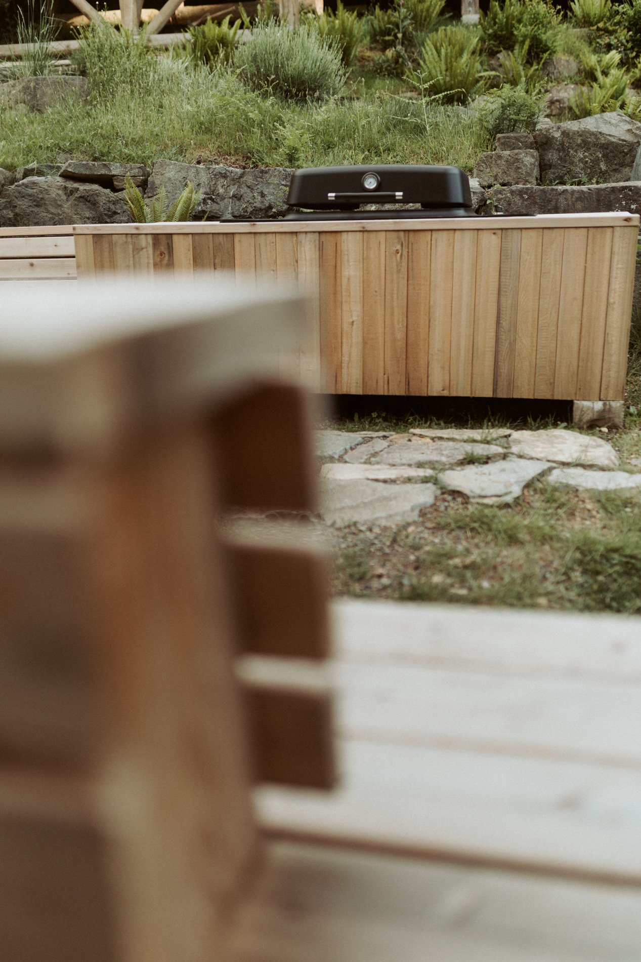 Close-up of a black barbecue grill on a stone patio with a wooden fence and greenery in the background.