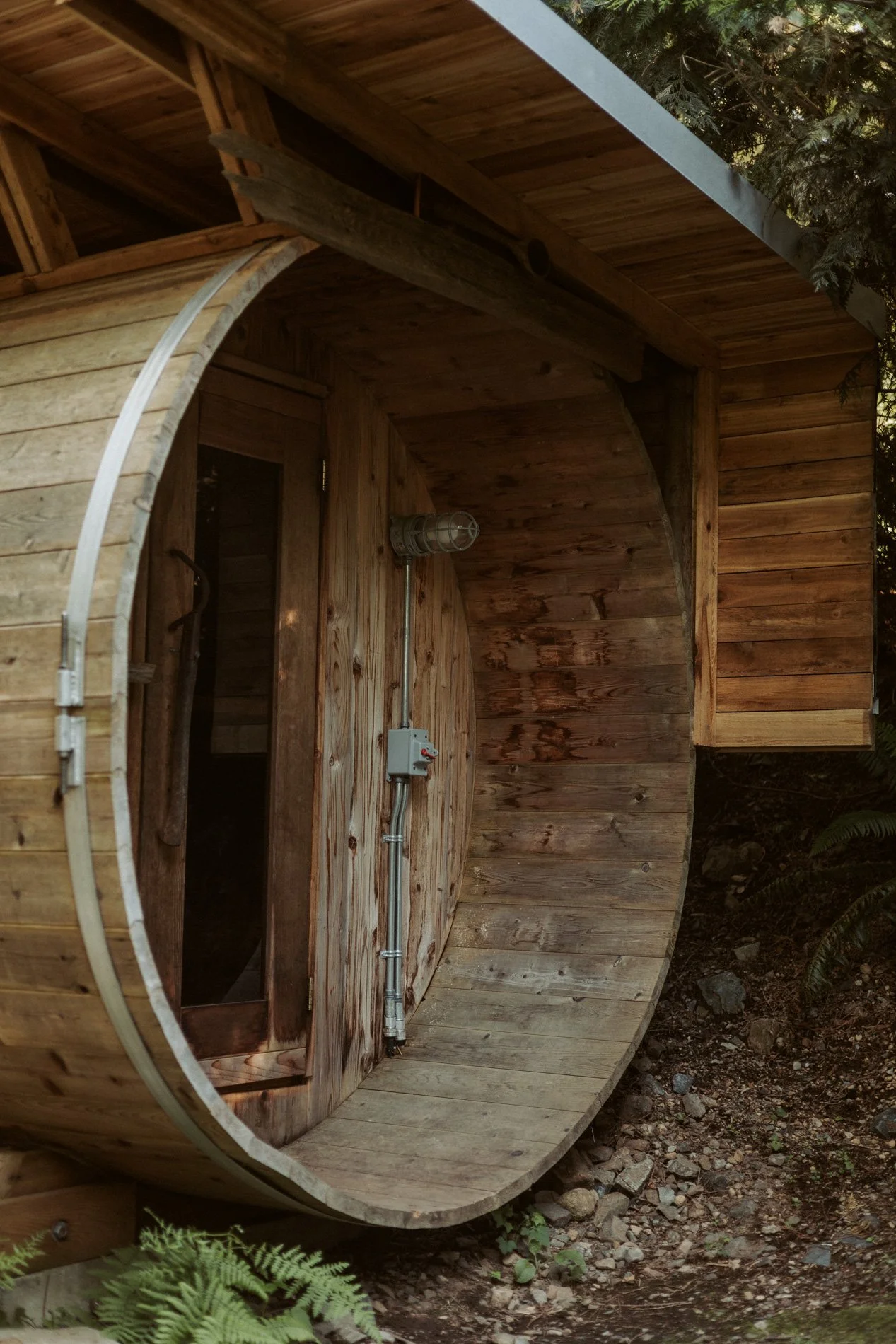 A small, round wooden sauna with a door and a window, surrounded by greenery and rocky ground.