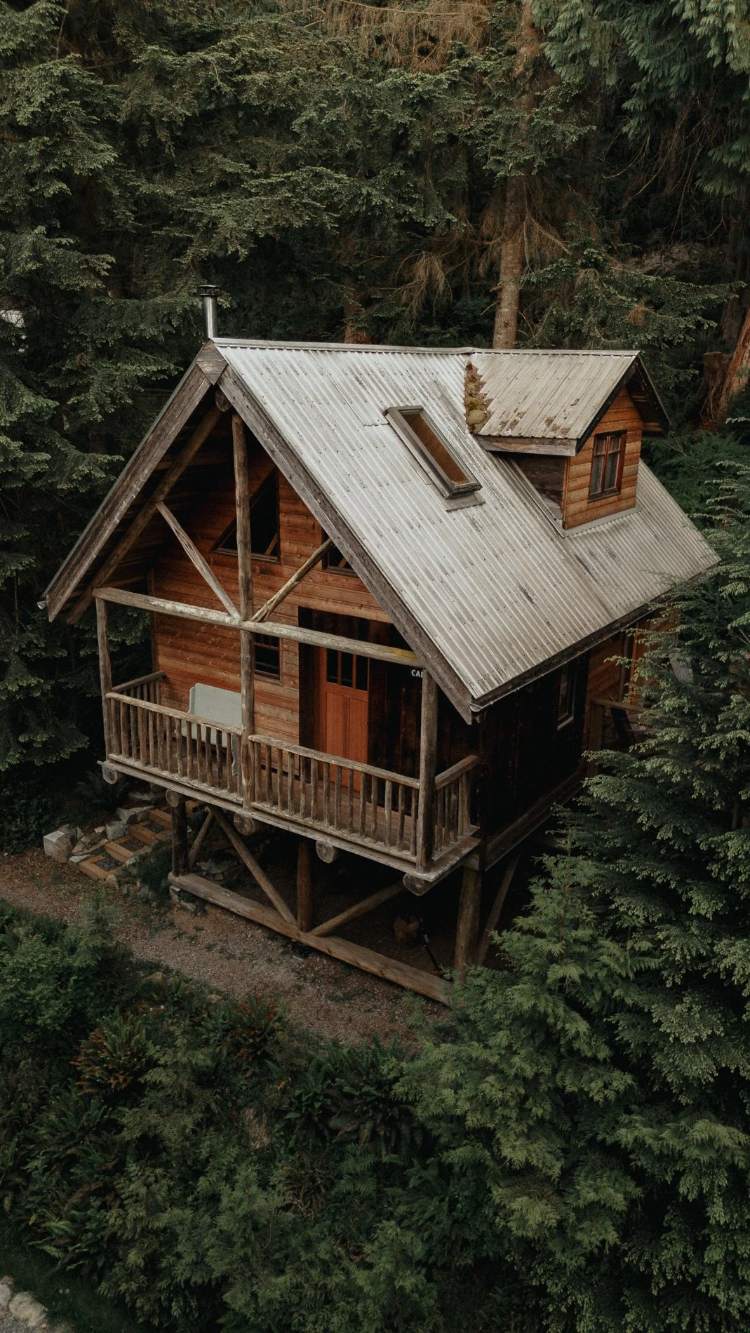 A wooden house with a metal roof, situated in a dense forest. The house has a small balcony and a skylight on the roof.