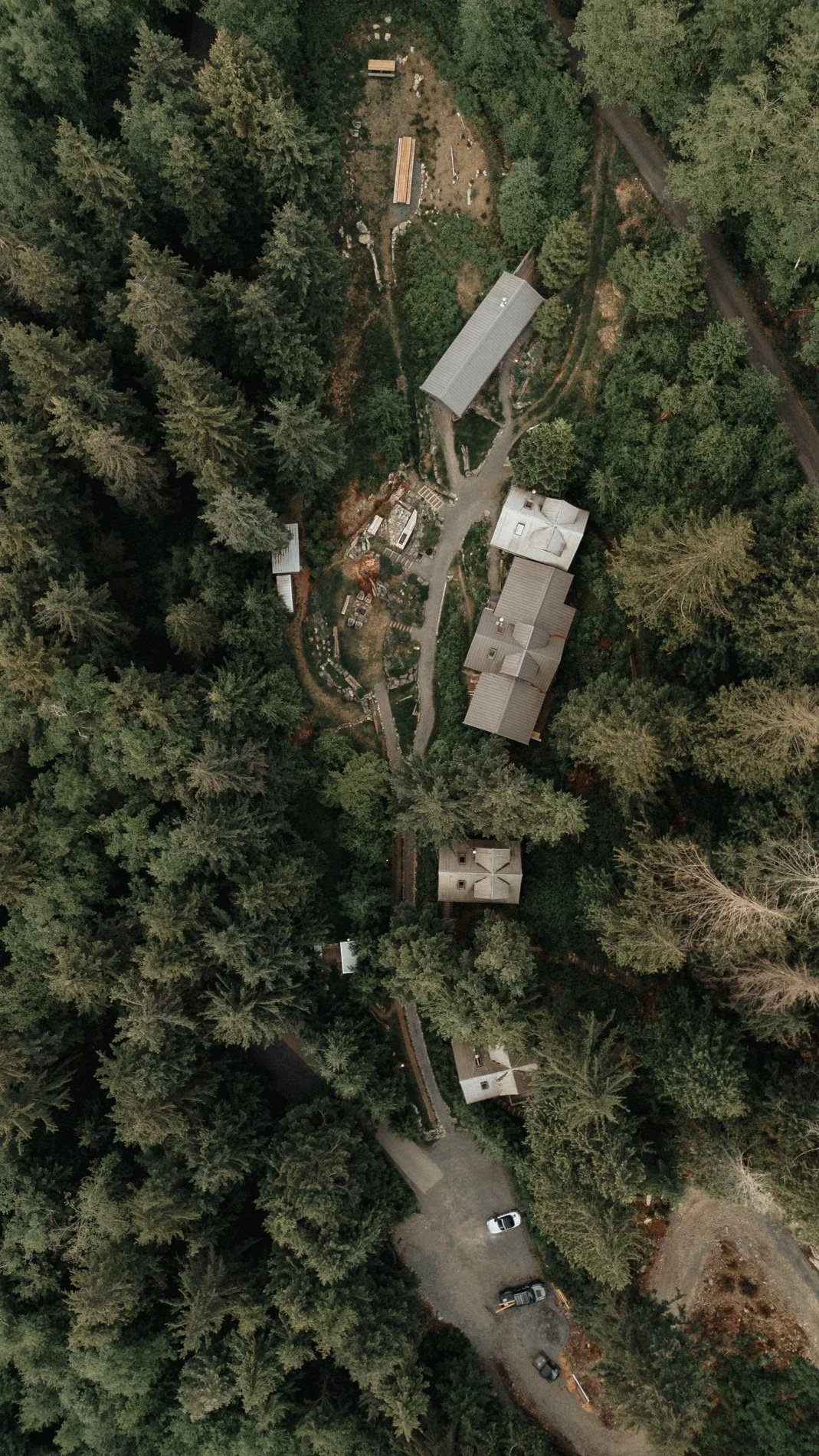 An aerial view of a property surrounded by dense trees, with multiple buildings, a winding driveway, and a parking area with several cars.