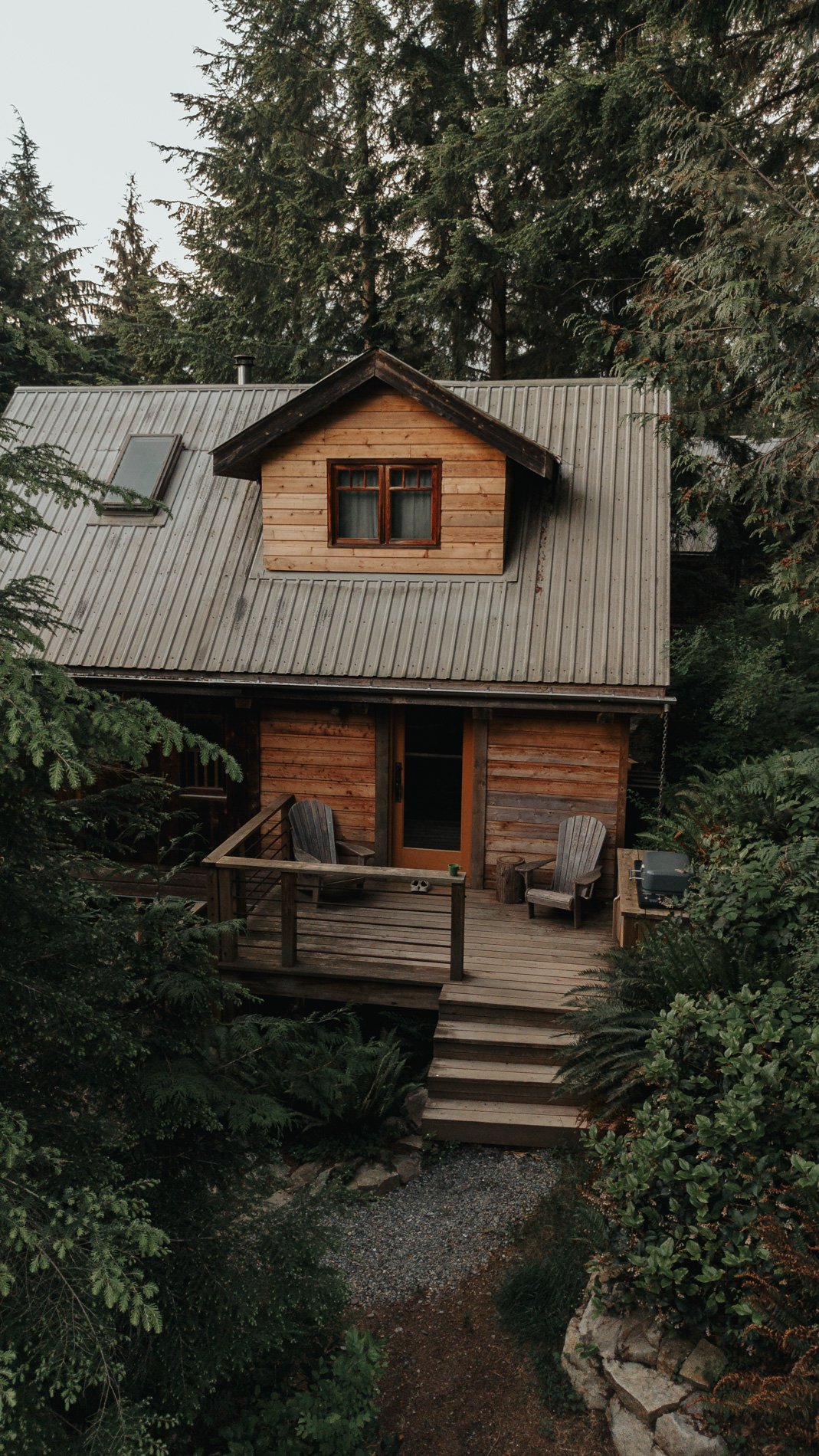 A wooden house with a gray metal roof, surrounded by tall trees and lush greenery, featuring a small balcony with two chairs.