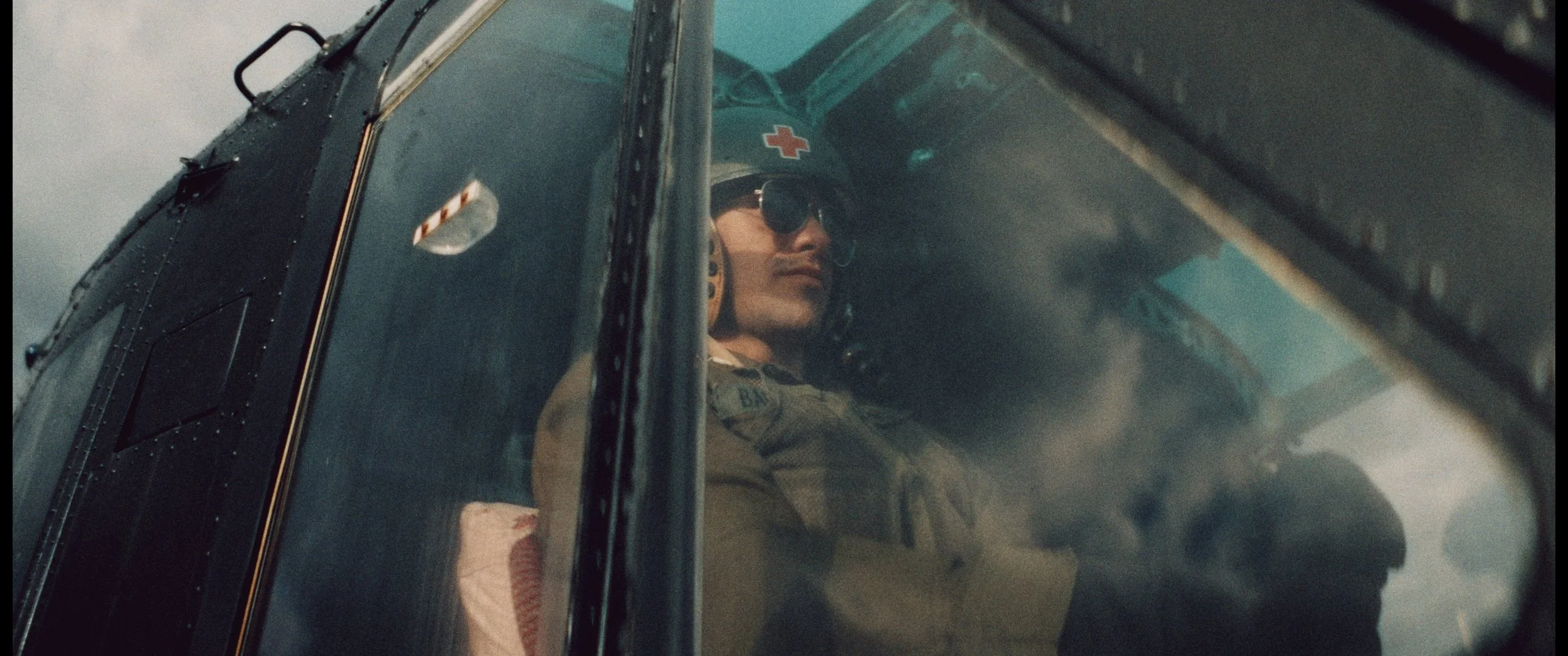 A male helicopter pilot inside the cockpit, wearing sunglasses and a helmet with a red cross symbol, looking out through the helicopter window.
