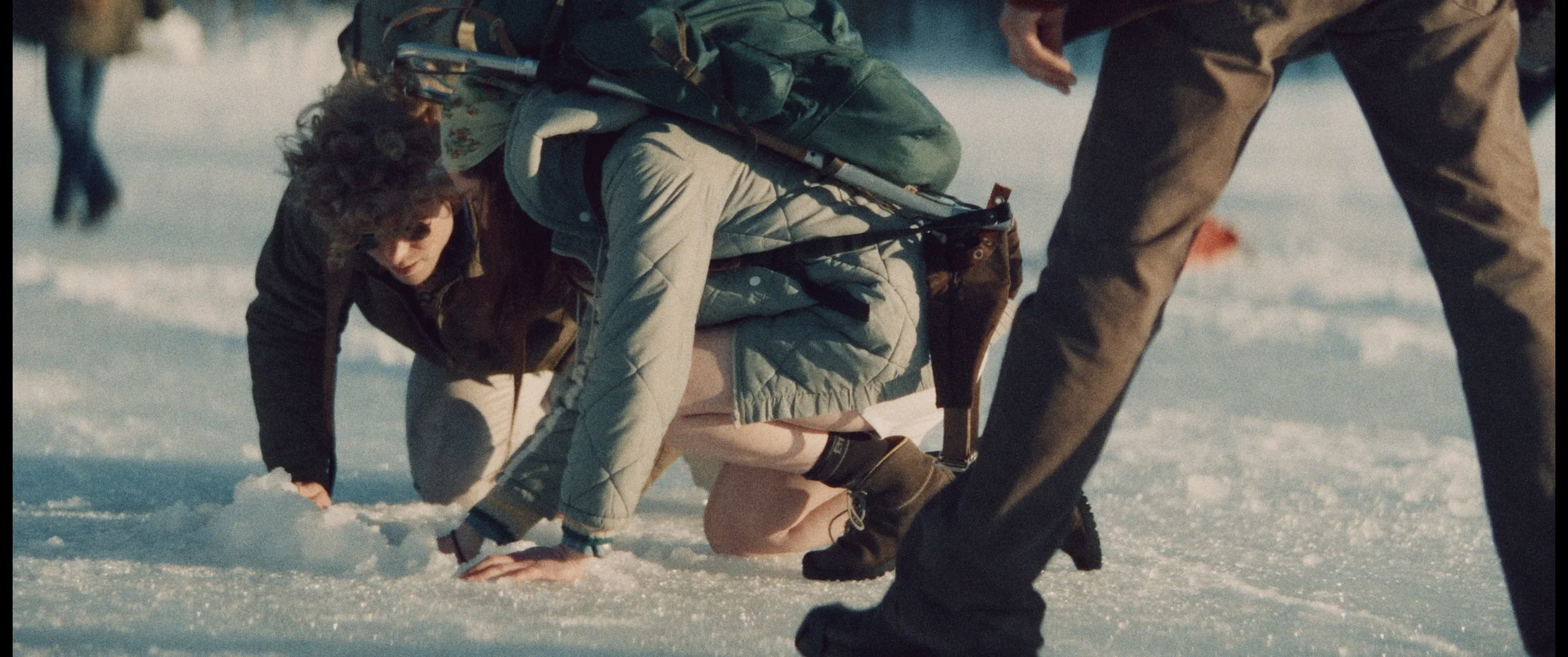 Person in winter clothing kneeling on snowy ground, looking distressed, with another person standing nearby. The scene appears to be outdoors in a cold environment.