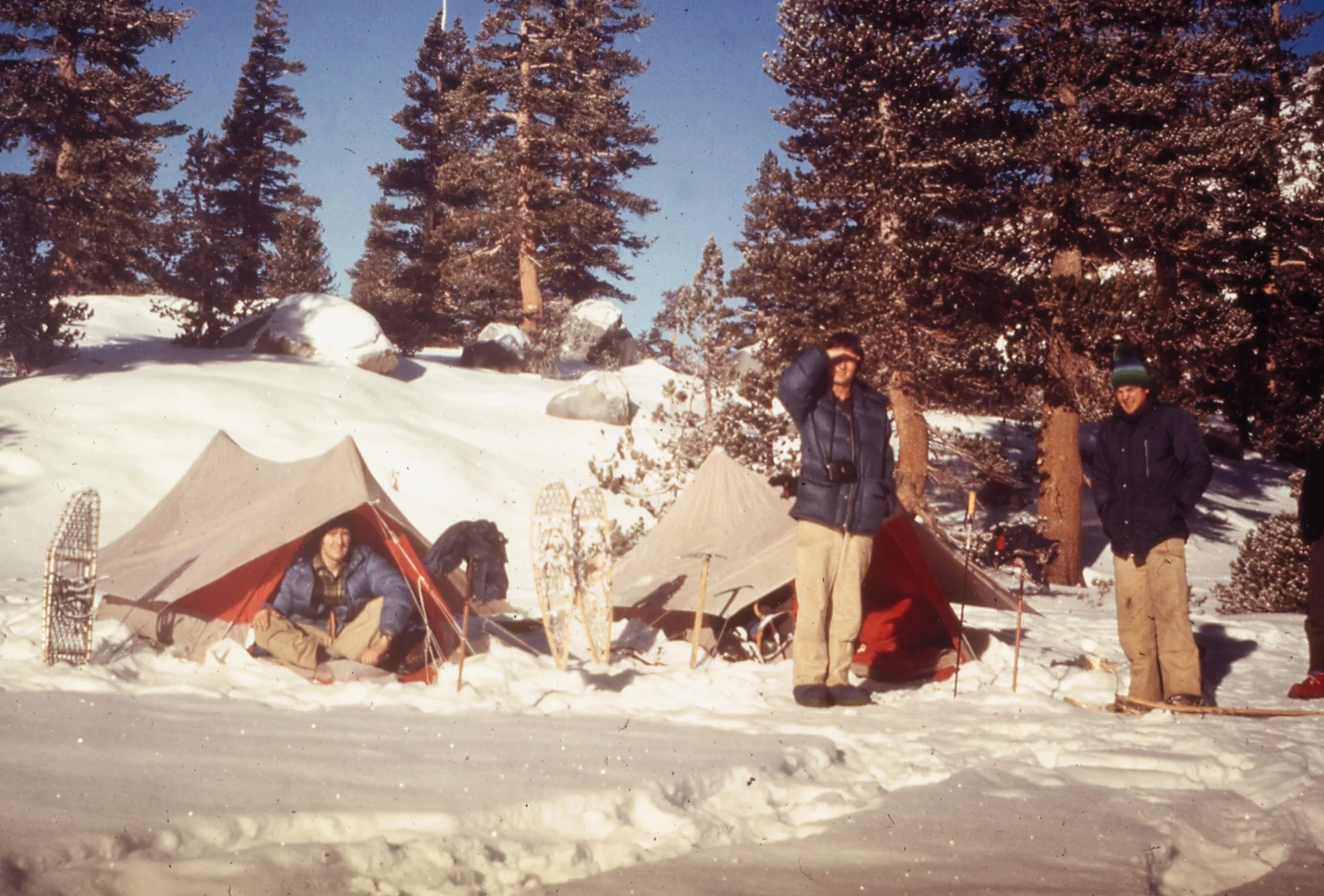 A snowy outdoor scene with three people, several tents, and evergreen trees in the background. Two people are standing, dressed in winter clothing, and one person is sitting near a tent.