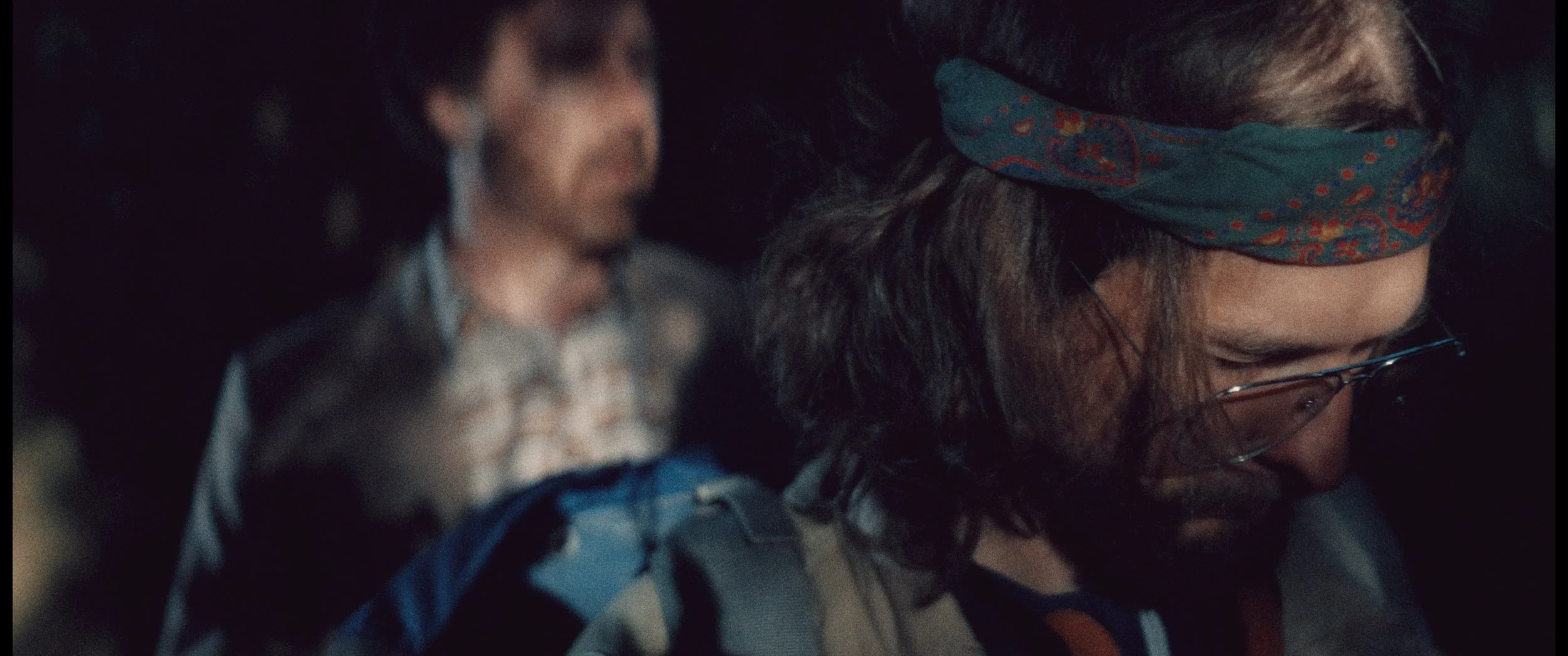 Close-up of a person wearing glasses and a patterned bandana, with another person blurred in the background.