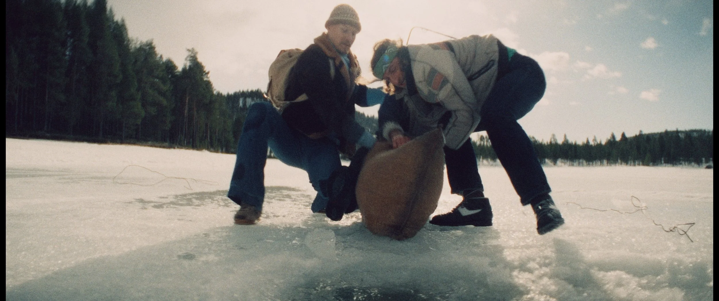 Two people in winter clothing on a snow-covered lake, leaning over and holding a brown bag, with a forest and blue sky with clouds in the background.