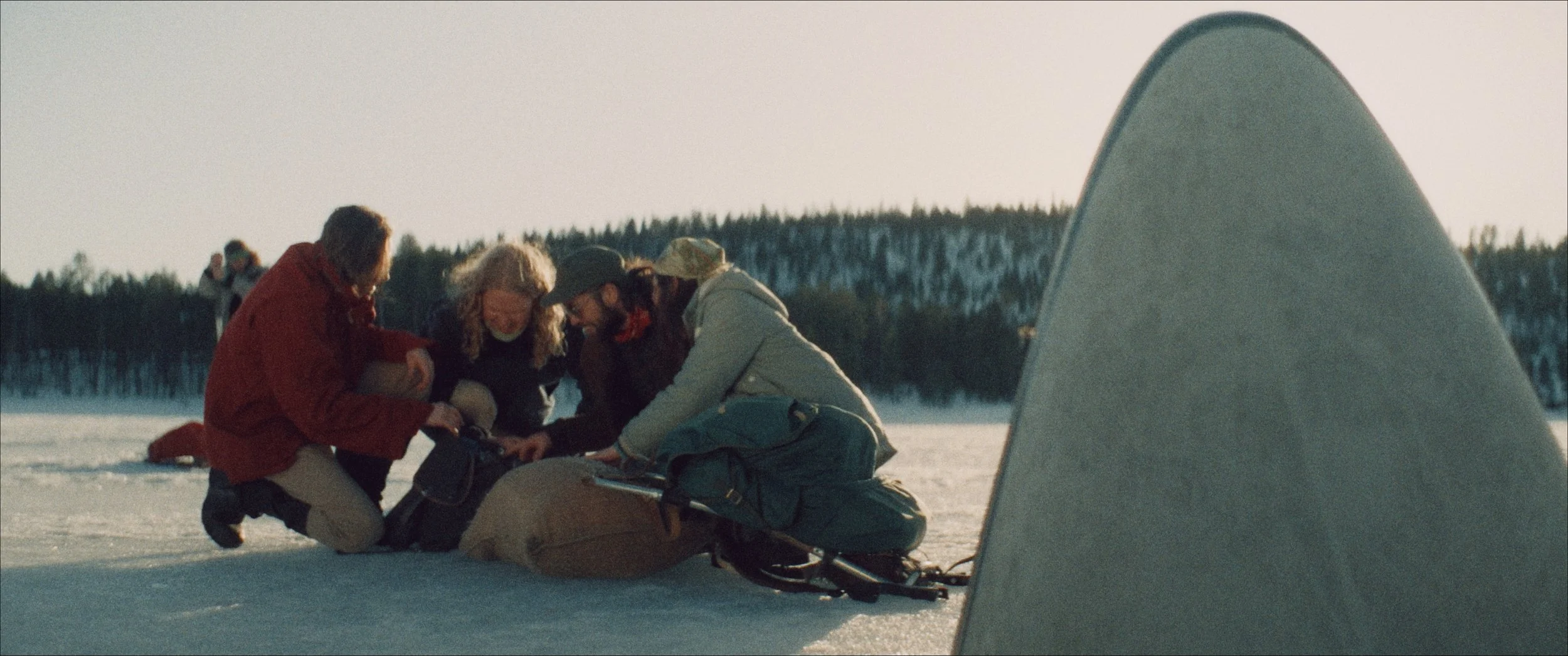 Four people helping a person who has fallen off a snowmobile on a snow-covered landscape with a forest in the background.
