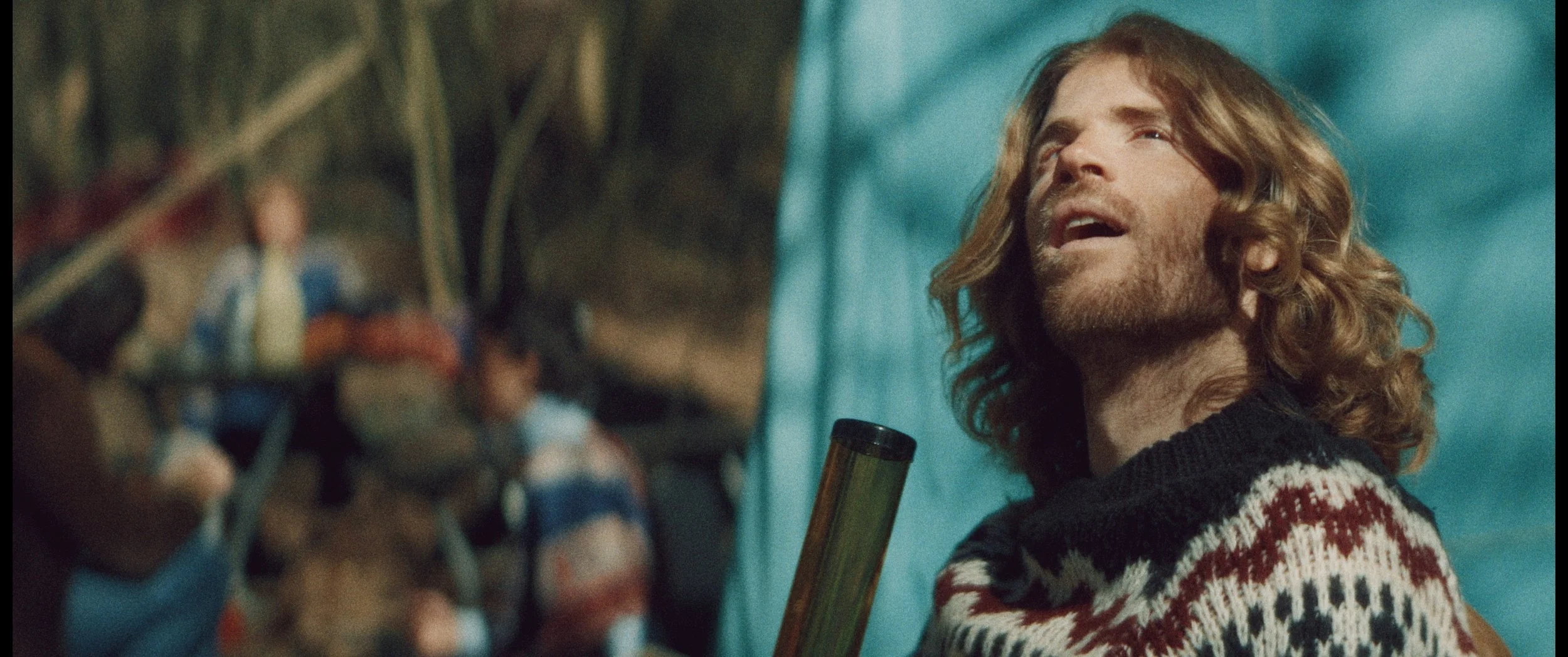 A man with long, curly hair and a beard, wearing a festive sweater, holds a guitar and singing outdoors during daytime, with a group of people in the background near trees.