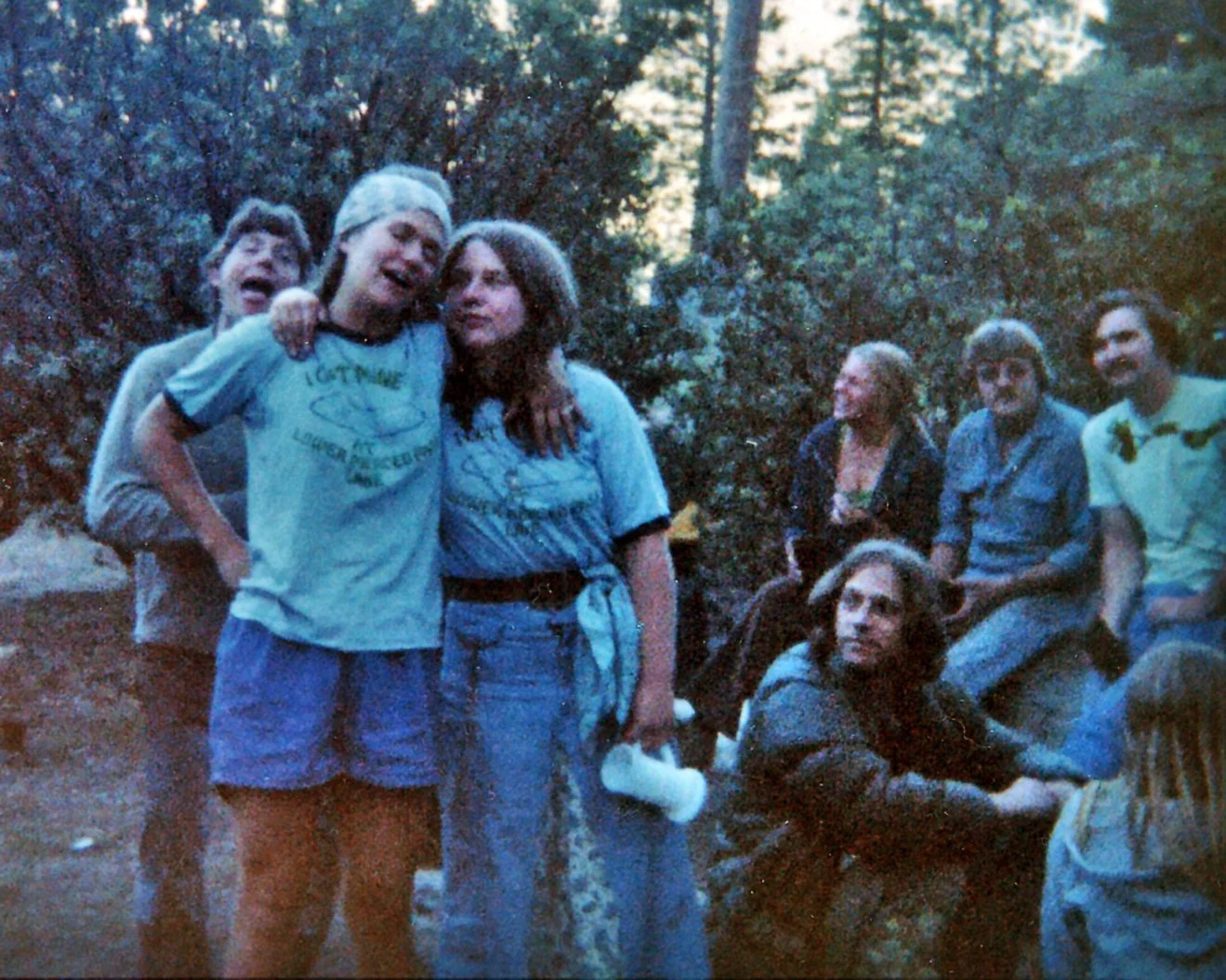 A group of seven people outdoors in a wooded area during evening. Two people are standing in the front, hugging and singing, with others sitting or standing behind them, some smiling and others looking contemplative.