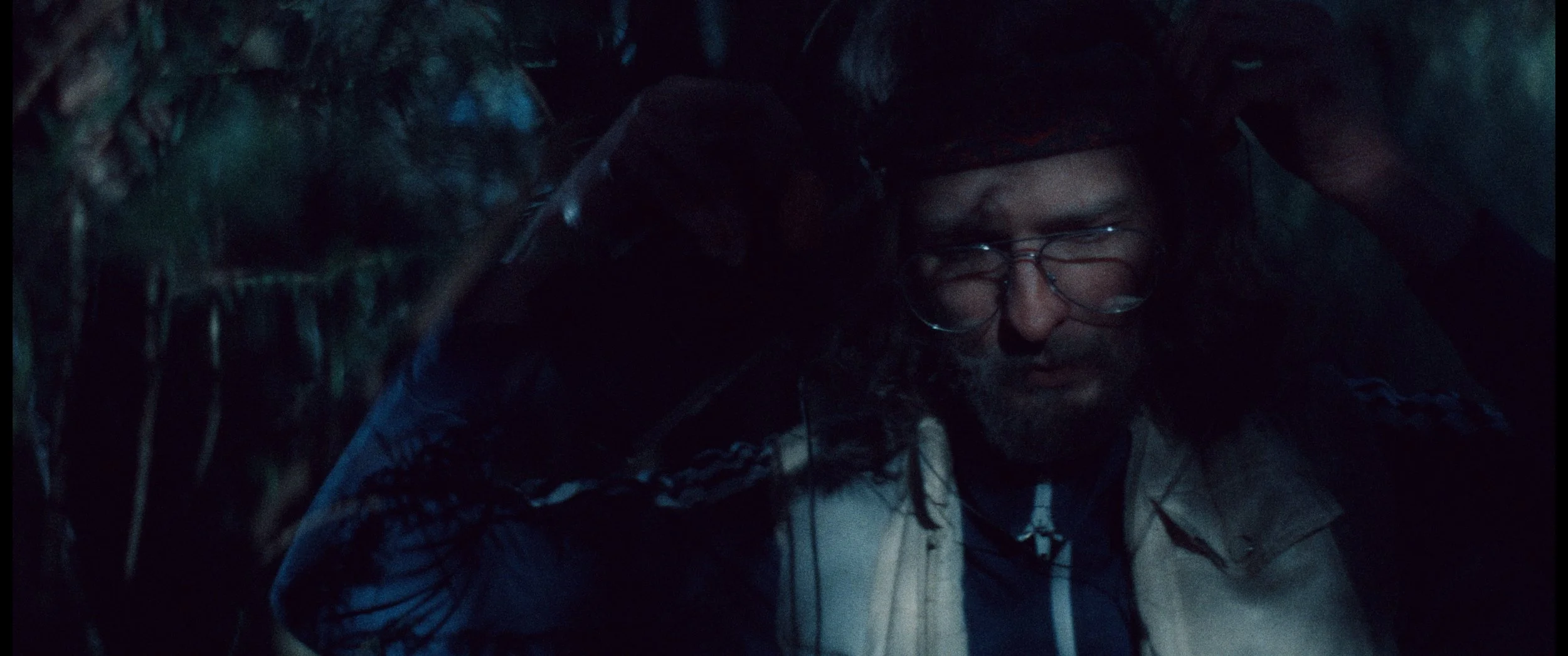 Person with glasses and curly hair in a dark outdoor environment, possibly in a wooded area at night.