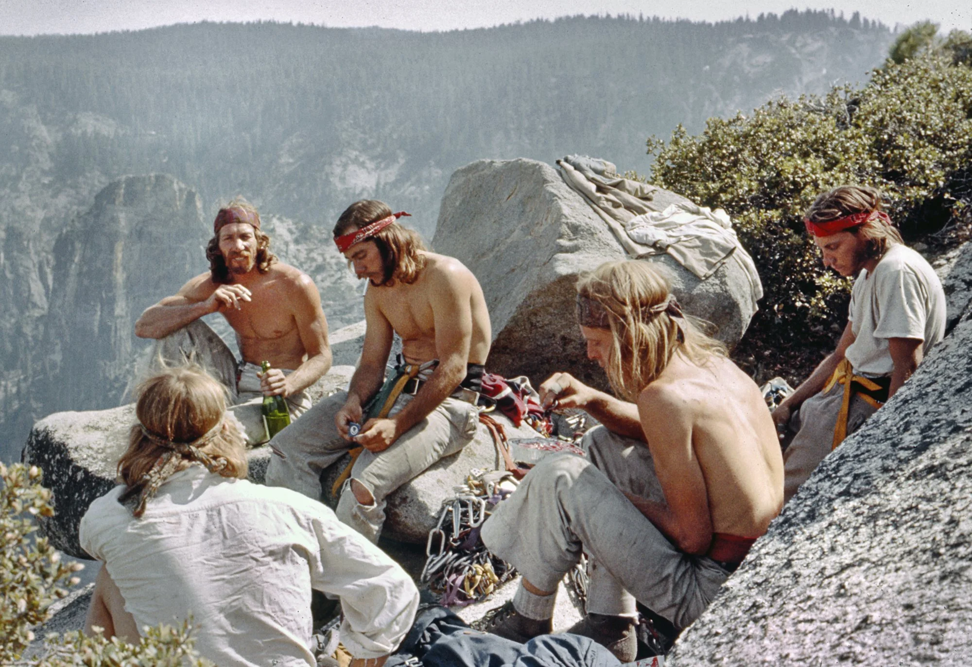 A group of young adults resting on rocks outdoors, surrounded by mountain scenery, some shirtless, wearing bandanas, and engaging in conversation or preparing climbing gear.