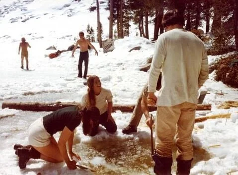 People in winter clothing gathered around a partially frozen snow-covered pond, some using shovels to clear ice, with snow and trees in the background.