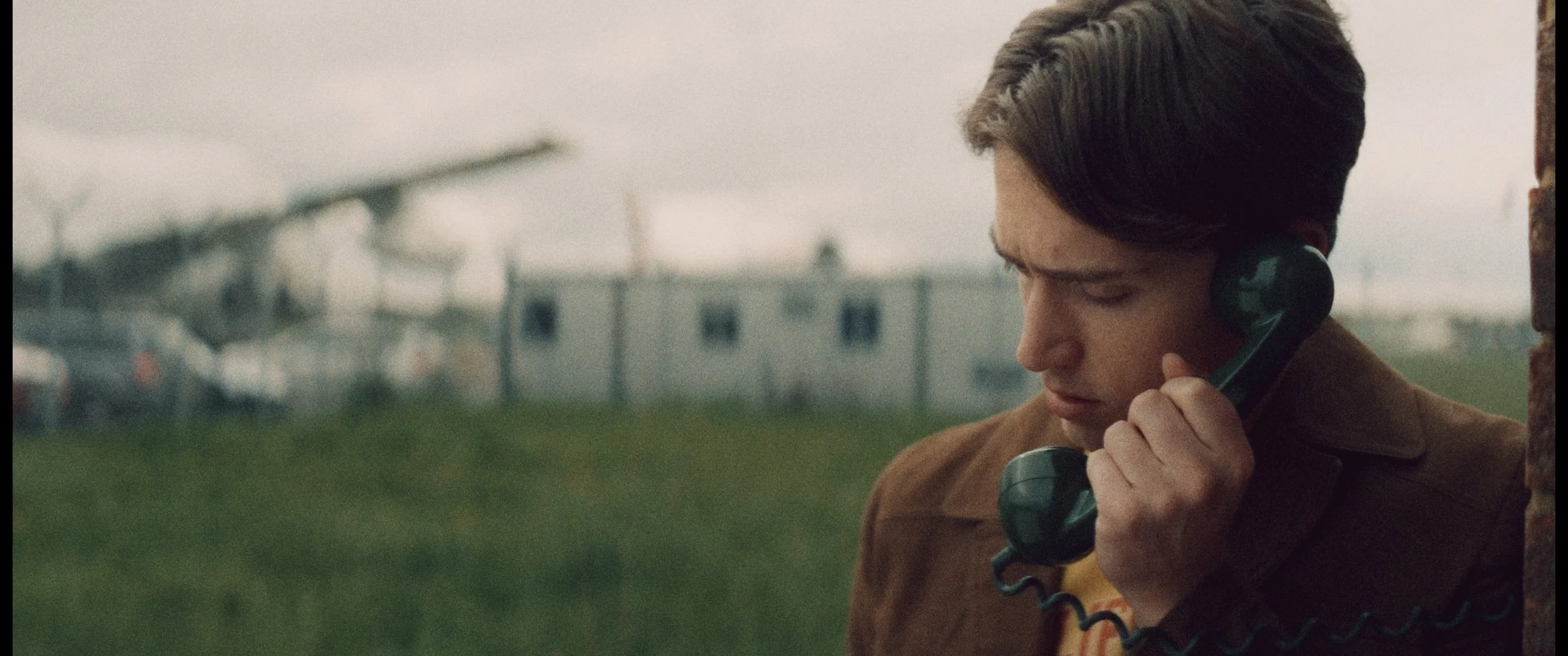 A young man with dark hair talking on a corded phone outdoors, with a blurred background of a field, wind turbines, and a trailer or mobile home.